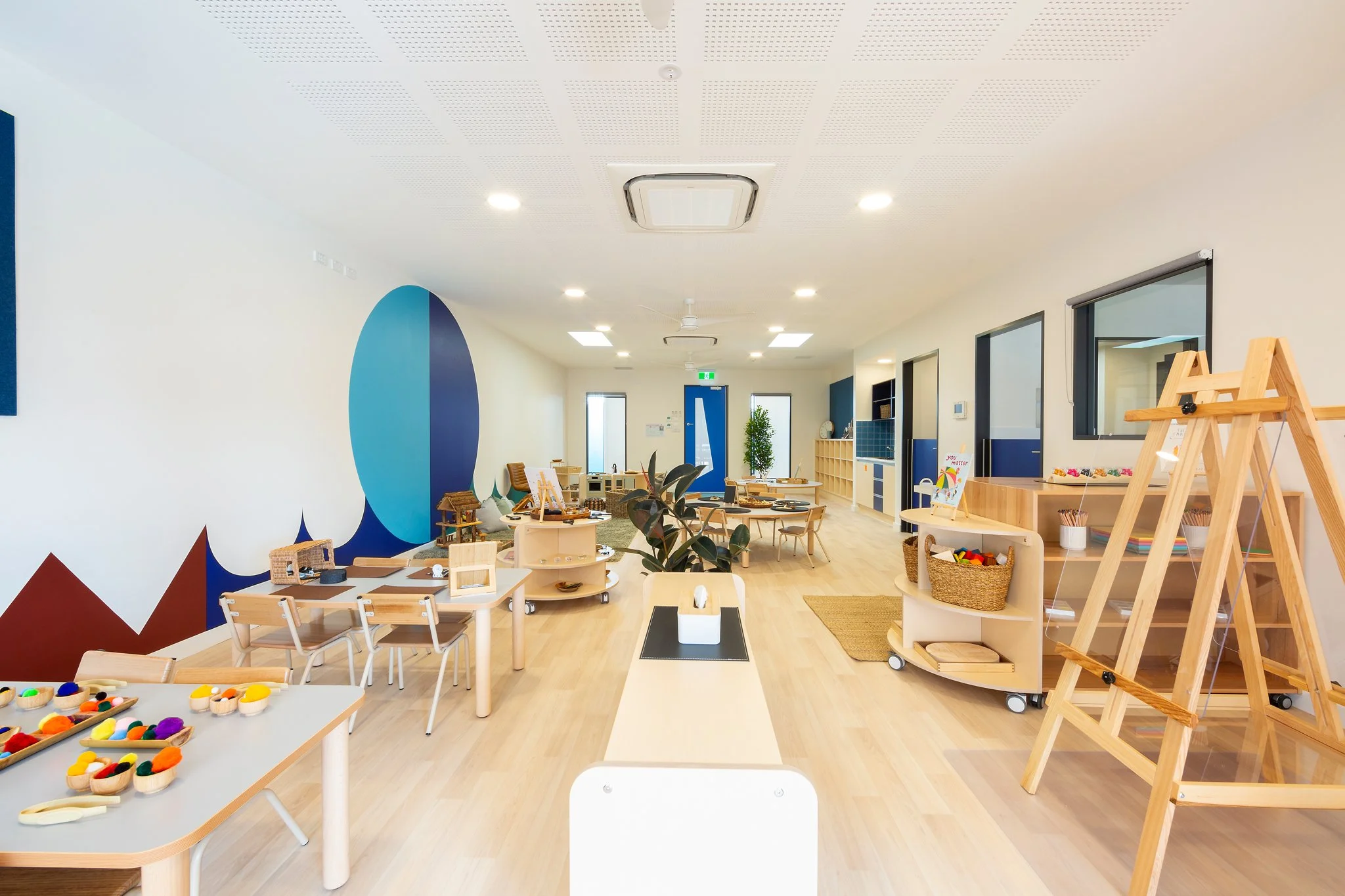 A spacious children's playroom with wooden tables and chairs, colorful toys, a blue and white wall mural, and large windows.