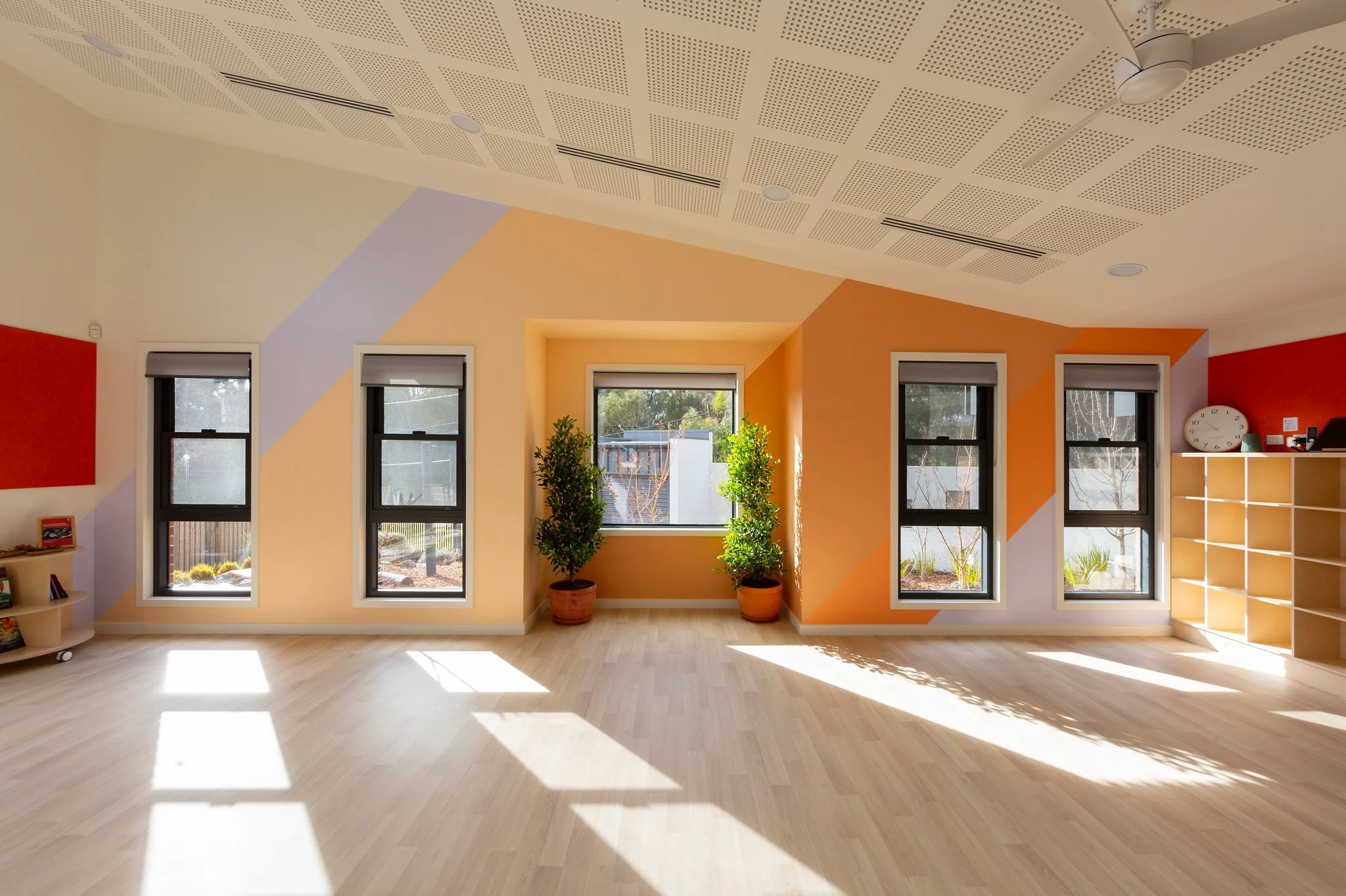 Bright room with large windows and colorful geometric wall design, potted plants, and wooden flooring.
