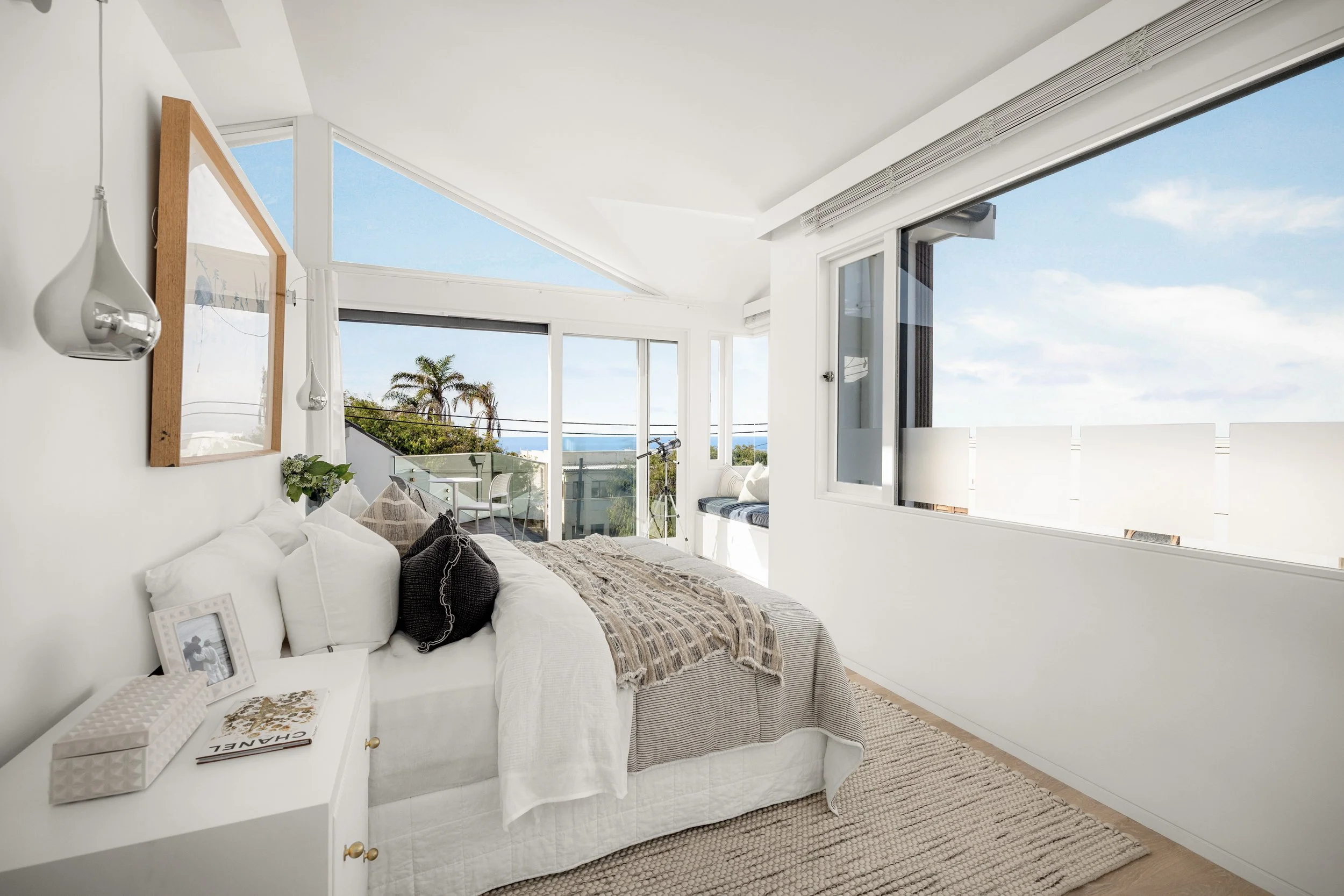 A bright, modern bedroom with large windows showing a view of a palm tree, the ocean, and a blue sky with clouds. The bed has white bedding, dark pillows, and a beige throw blanket. A white nightstand holds a photo frame, a book, and a tissue box. Th