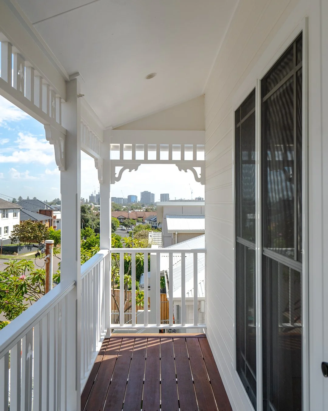 View from a balcony with white railing and wooden floor, overlooking a neighborhood with trees, houses, and city buildings in the distance under a partly cloudy sky.