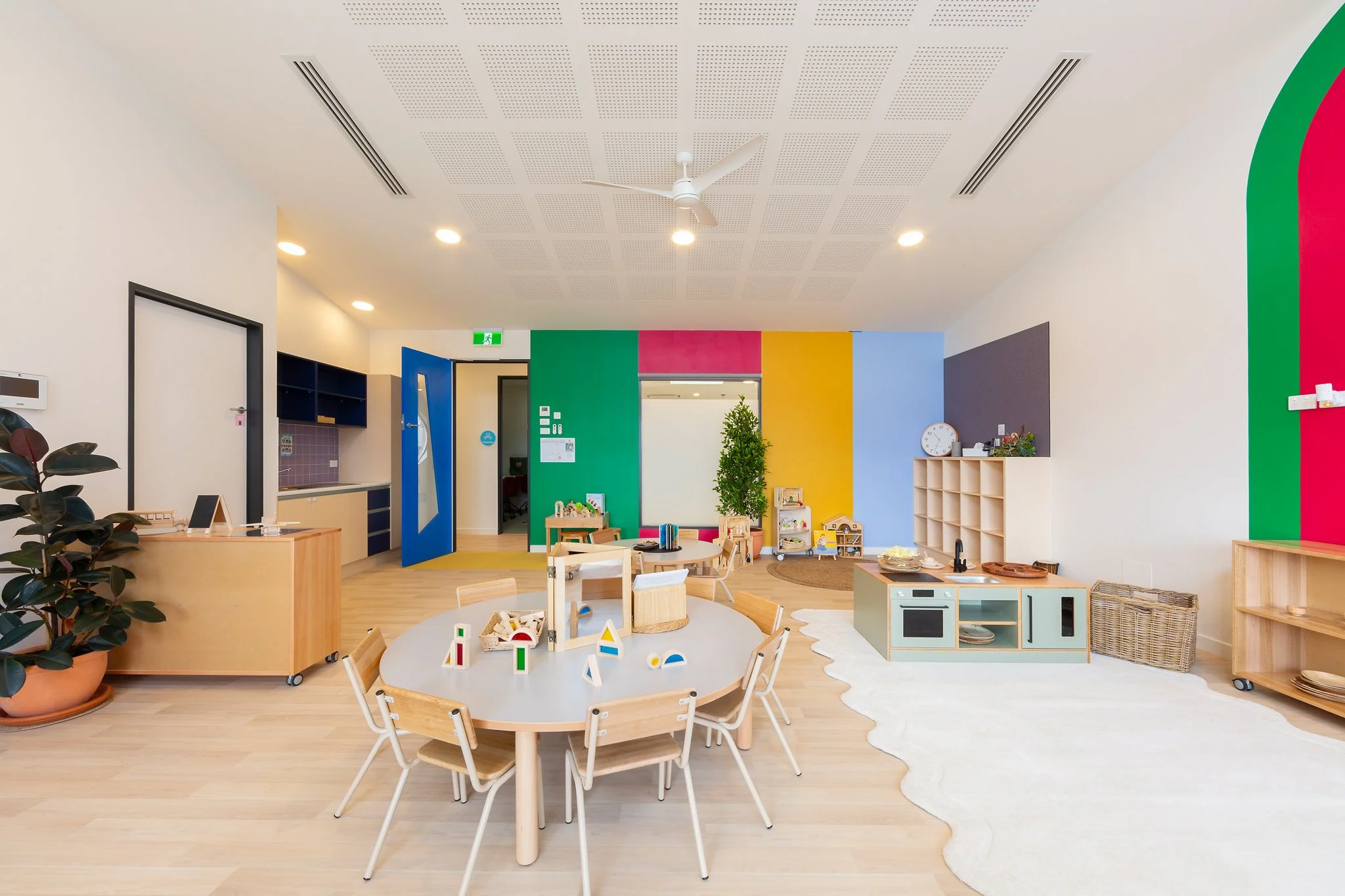 Colorful children’s playroom with a round table and wooden chairs, toys, plants, a white rug, and bright painted walls in green, pink, yellow, and purple. Ceiling has a fan and lighting, with a small kitchenette area and storage.