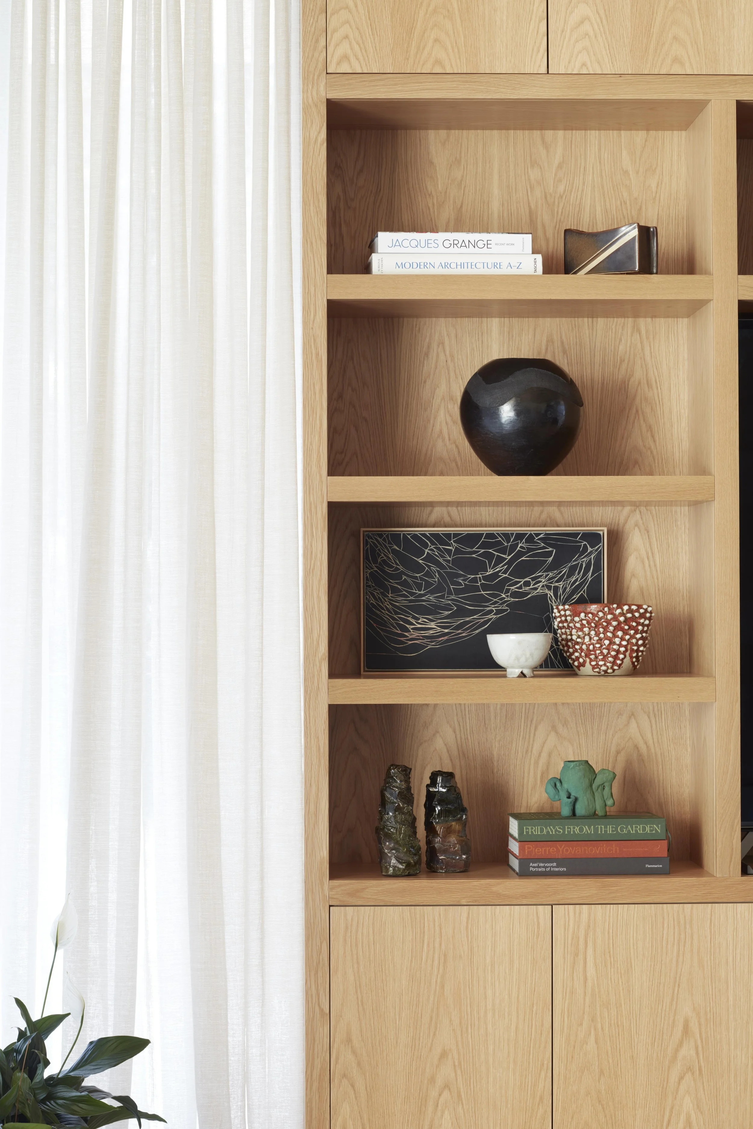 A wooden bookshelf with decorative items and books, beside a white curtain and a green plant.