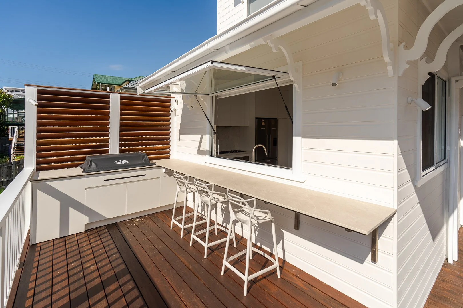 Outdoor balcony with a built-in grill, bar-height counter with four white chairs, white siding, window with an awning, privacy wooden fence, and a blue sky.