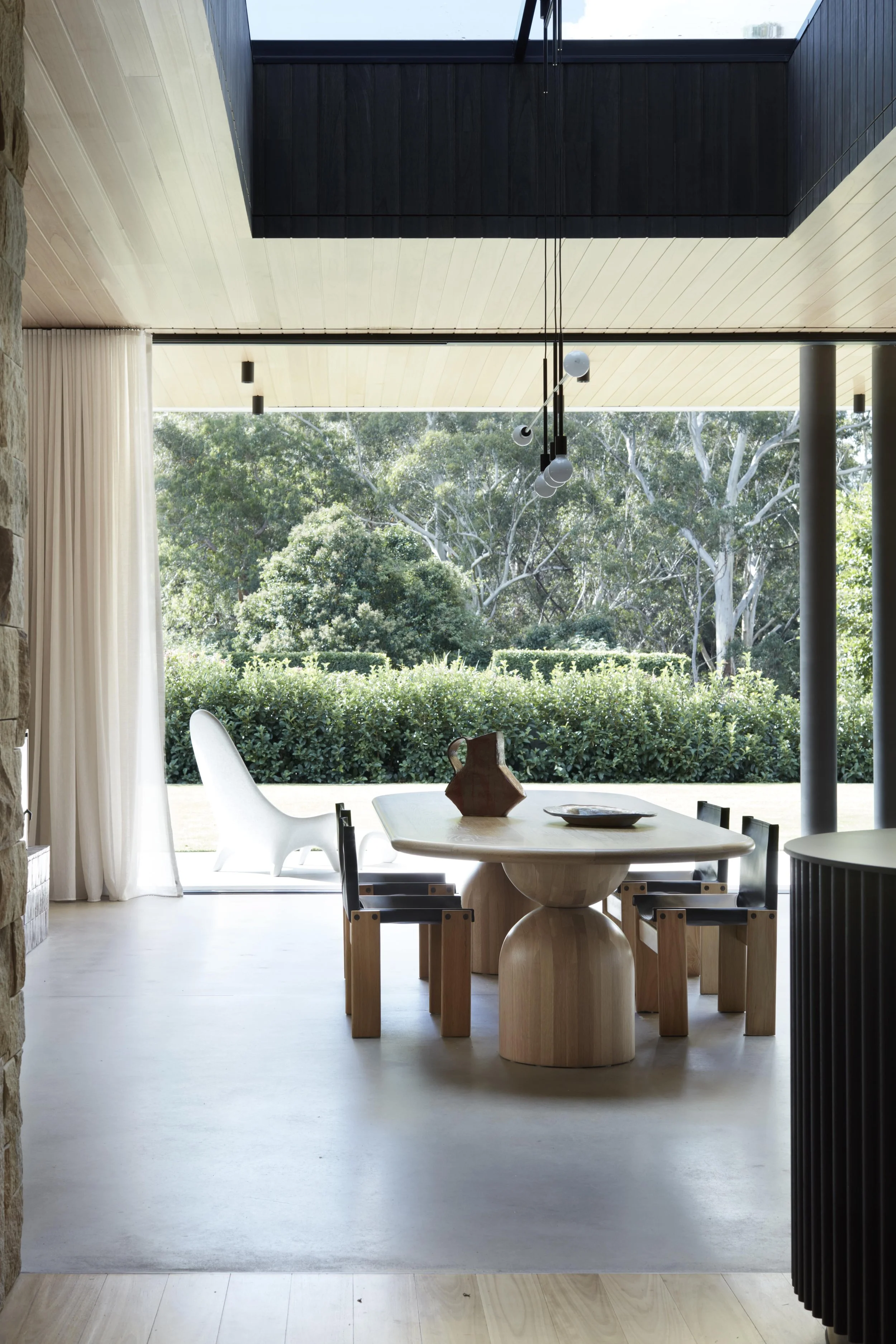 A modern dining area with a wooden table, black chairs, and decorative objects, opening to an outdoor view of lush green trees through large glass doors.