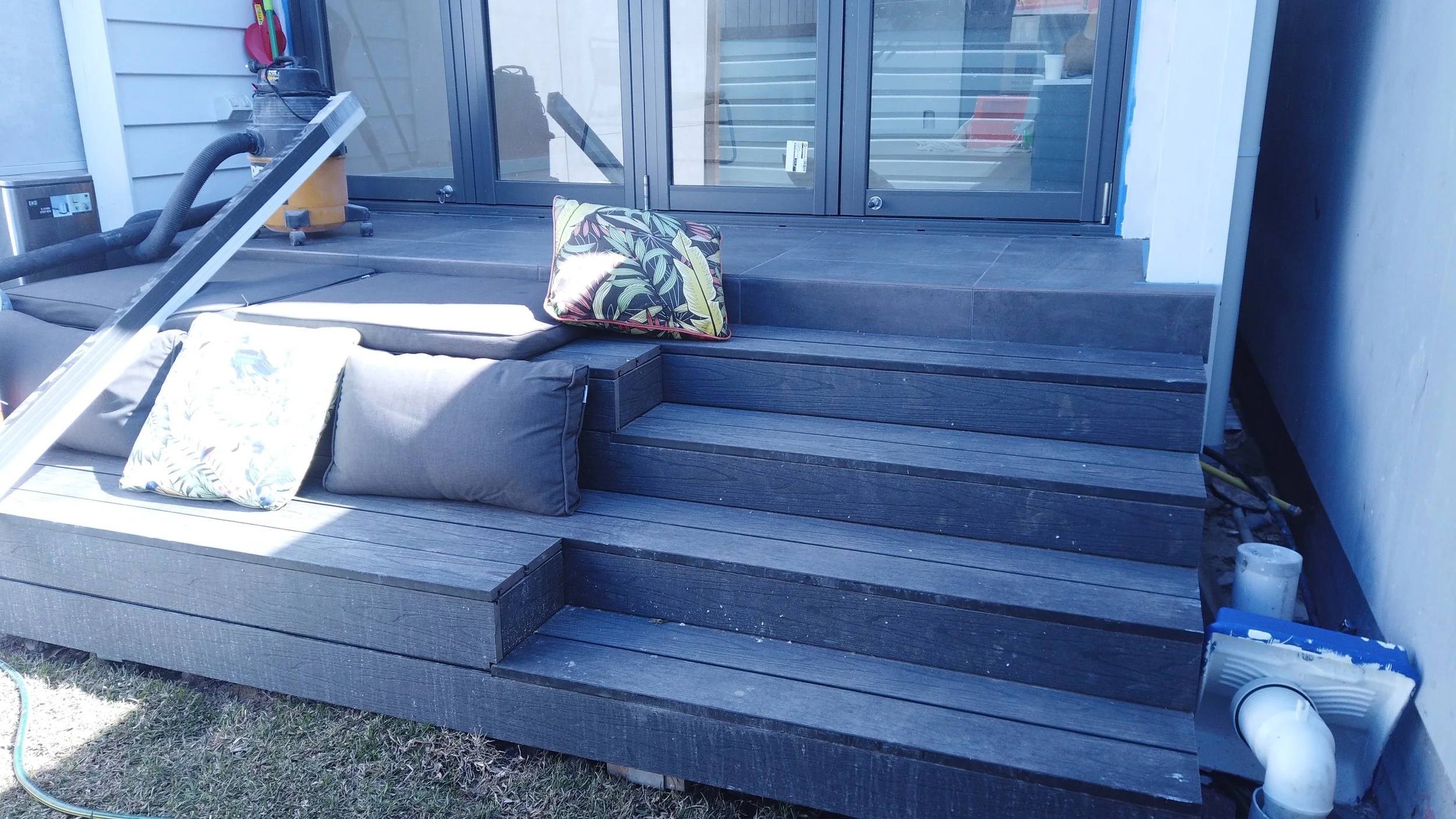 A backyard patio with dark wood steps leading to a sliding glass door, decorated with pillows, and some cleaning equipment and supplies nearby.