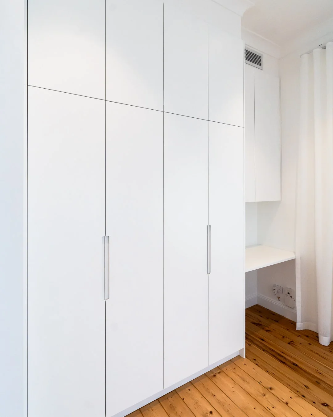 White built-in wardrobe with vertical handles in a room with wooden floor and a small desk area.