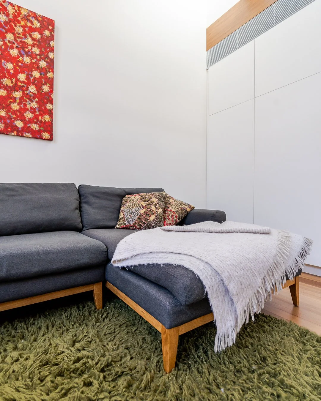 Living room corner with a gray sofa, patterned pillows, a white fringed throw blanket, green shaggy rug, white wall, red floral painting, and white cabinetry with a wooden trim.