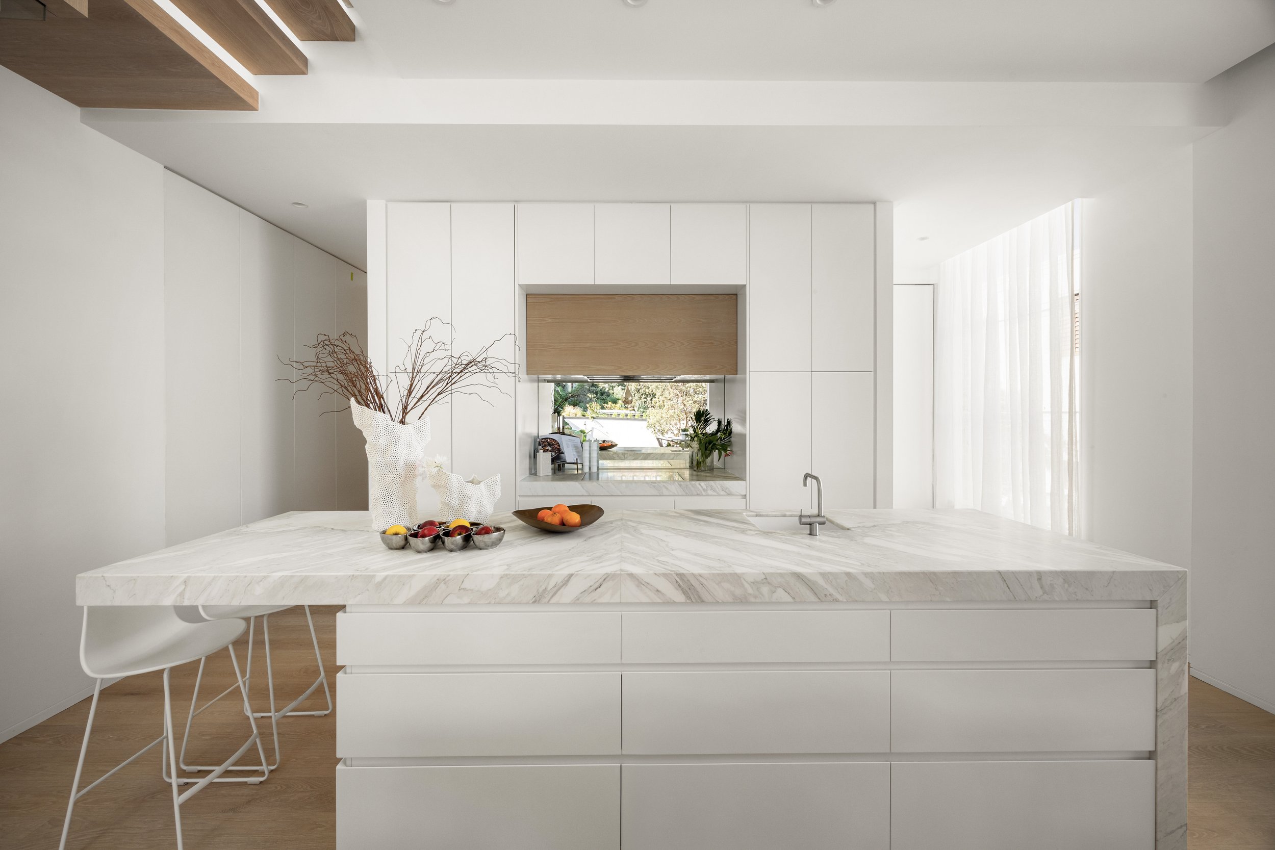 Modern white kitchen with a marble island, decorative vases, and bowls of fruit, bright natural light through sheer curtains.