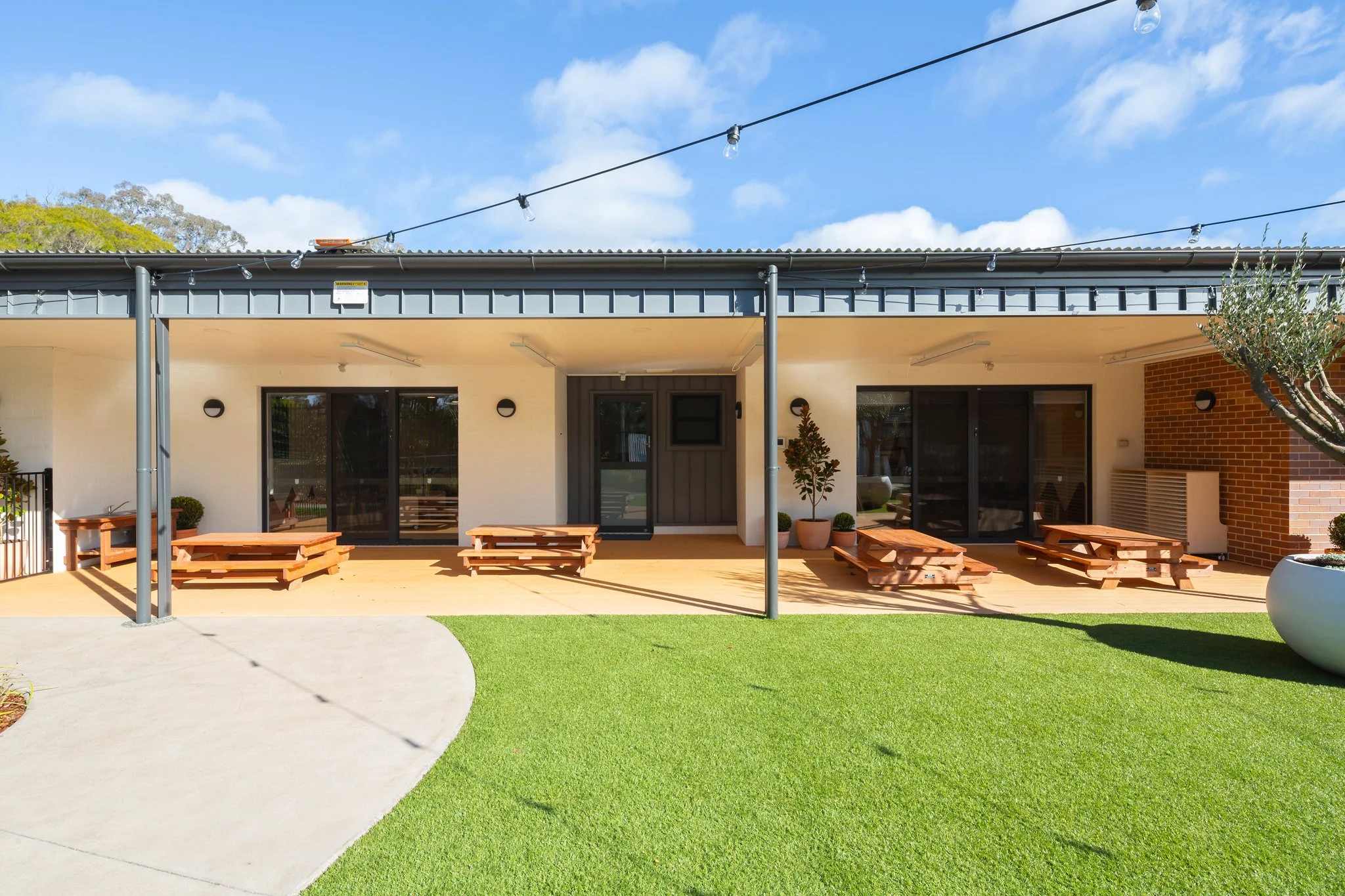 Outdoor patio area with wooden picnic tables, potted plants, and a building with sliding glass doors, string lights, and a brick chimney under a blue sky with clouds.