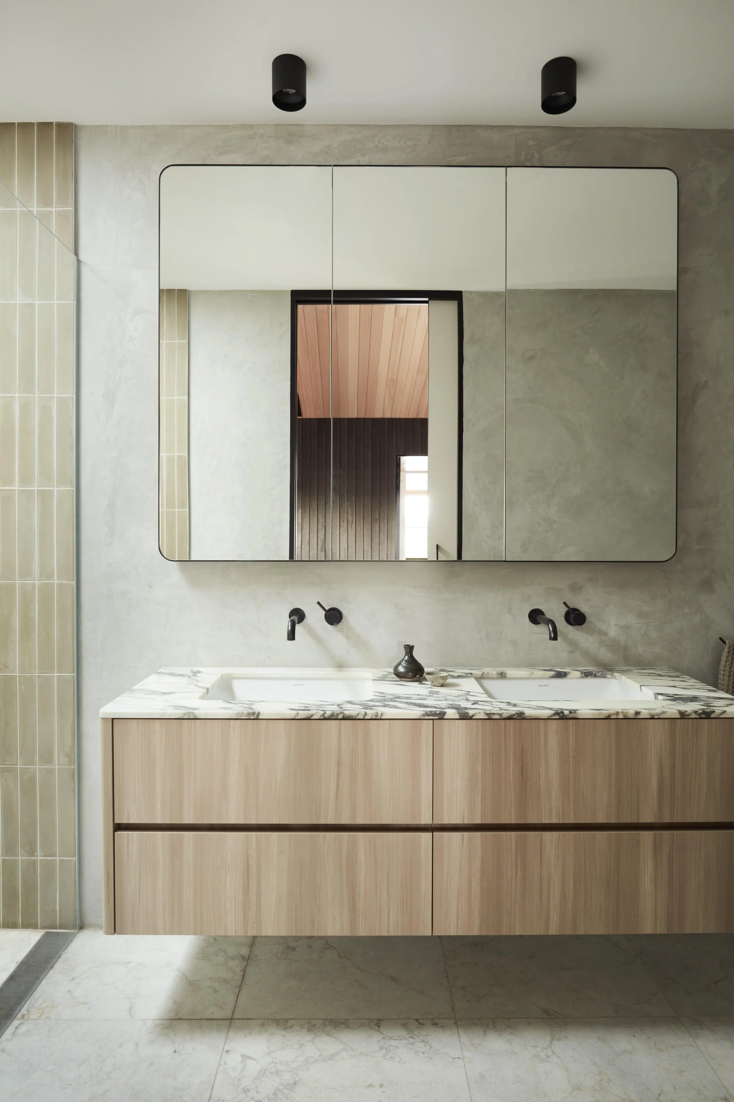 Modern bathroom vanity with a marble countertop, light wood cabinets, and two undermount sinks. A large mirror with rounded corners reflects a wooden ceiling and window. Two black wall-mounted faucets are above the sinks, and a small decorative objec