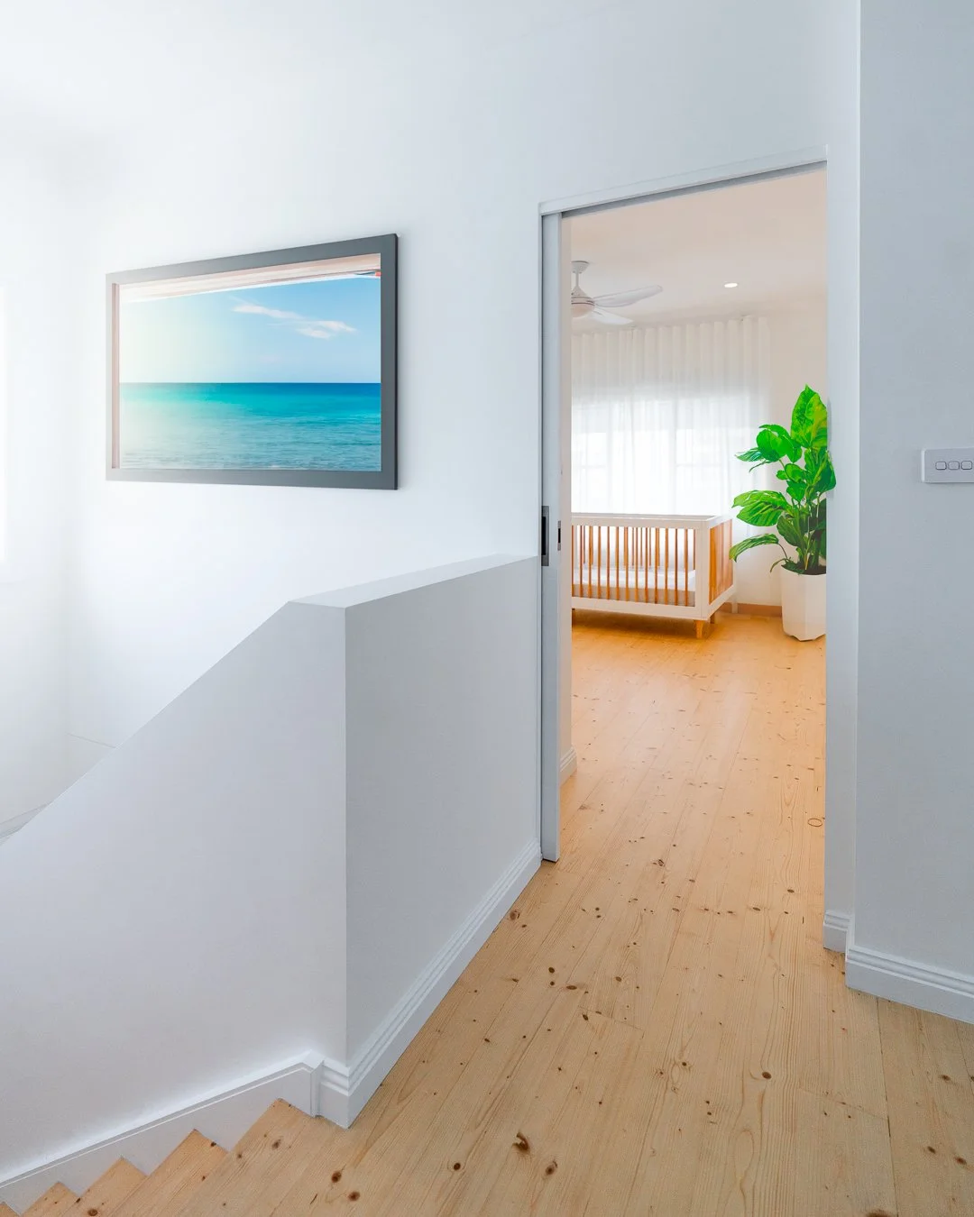 Interior view of a modern home with white walls, light wood flooring, a framed beach scene photo on the wall, a room with a crib and large green potted plant, and sheer curtains letting in natural light.