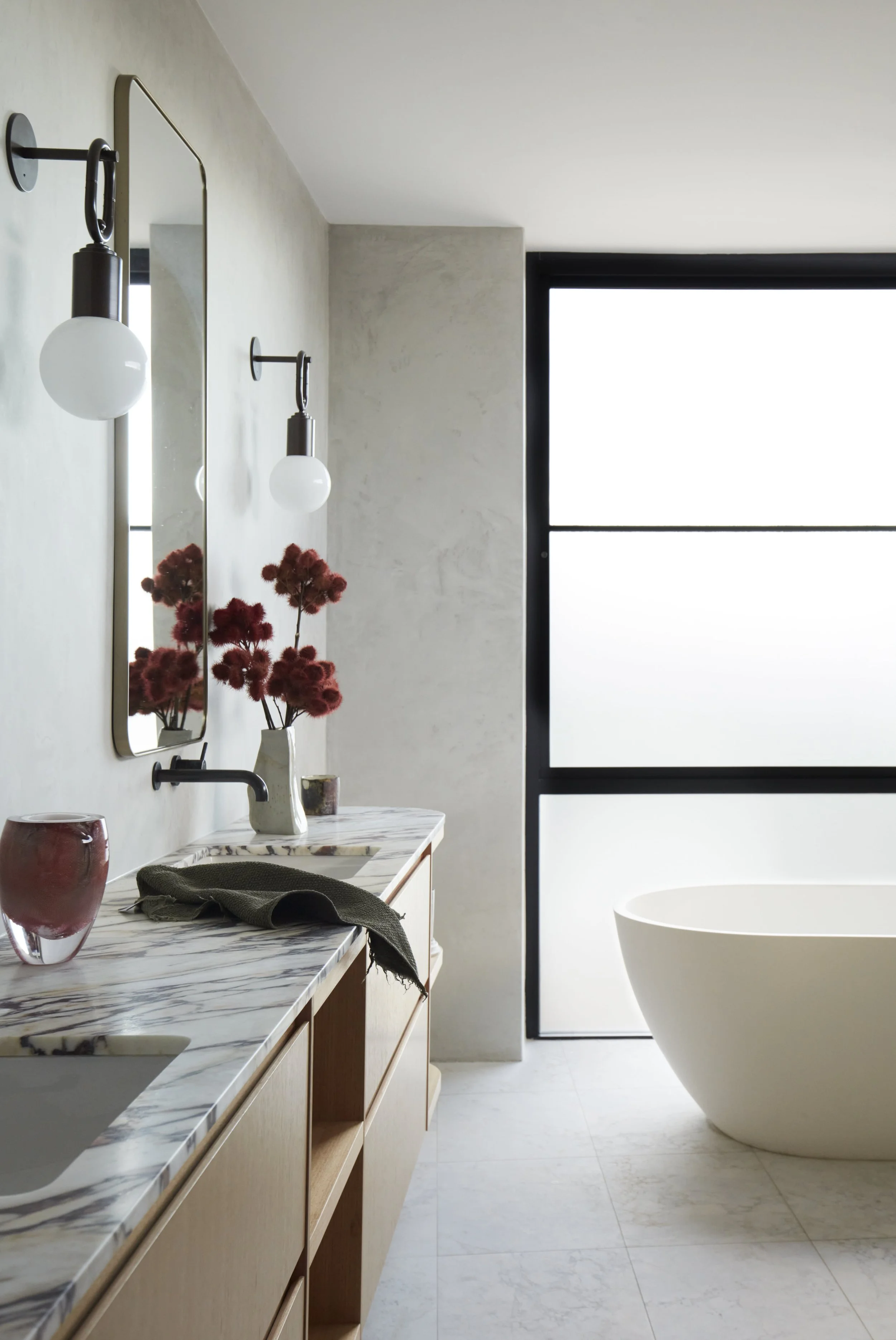 Modern bathroom with a white freestanding bathtub, wooden vanity with marble countertop, vase with red flowers, red and gray cups, black towel, wall-mounted mirror, black wall-mounted light fixtures, large black-framed window with frosted glass.