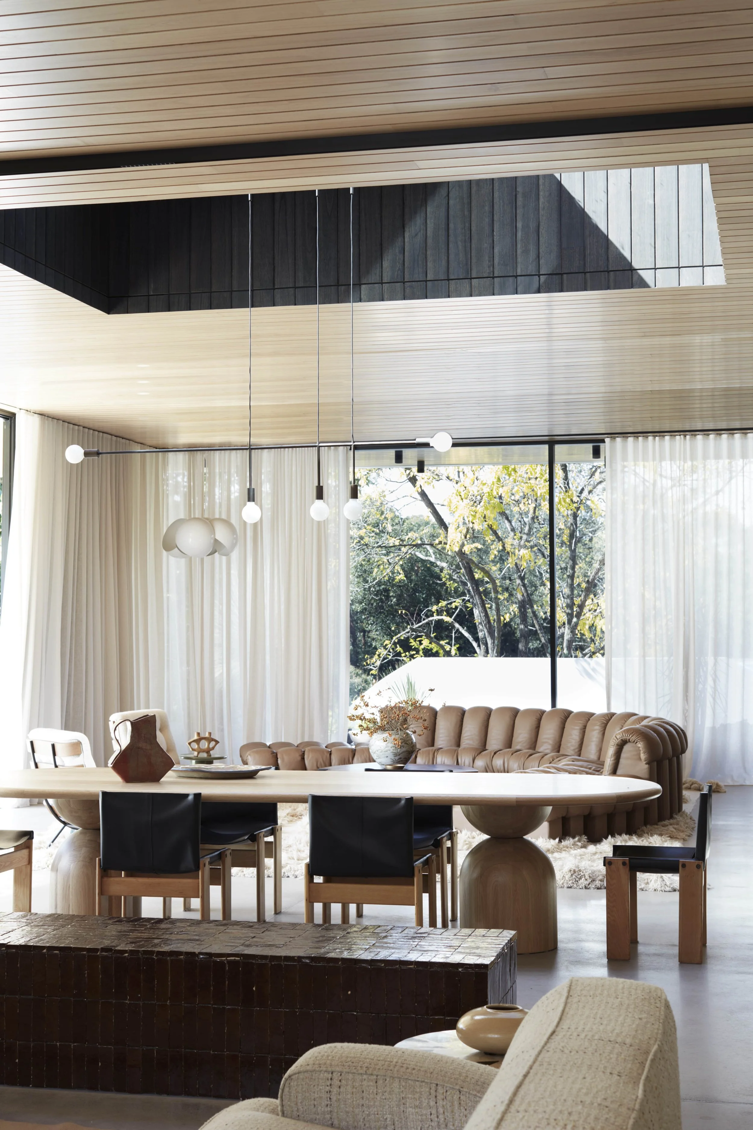 Modern living room with beige tufted sectional sofa, black chairs around a wooden dining table, large windows with white curtains, a shaggy rug, and a view of trees outside. Stylish lighting fixtures hanging from a wooden ceiling.