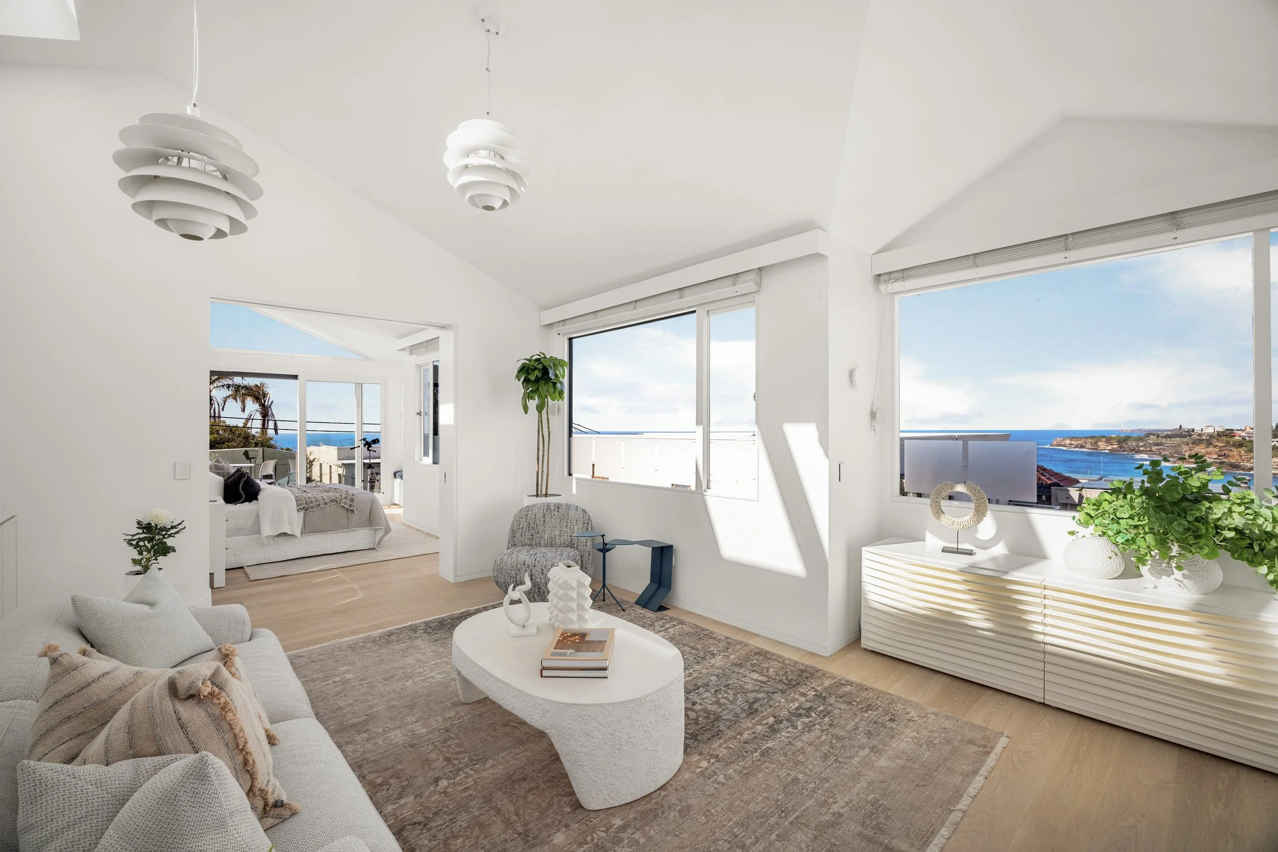 Bright living room with large windows showing blue sky and ocean view, modern white furniture, decorative plants, and minimalist artwork.