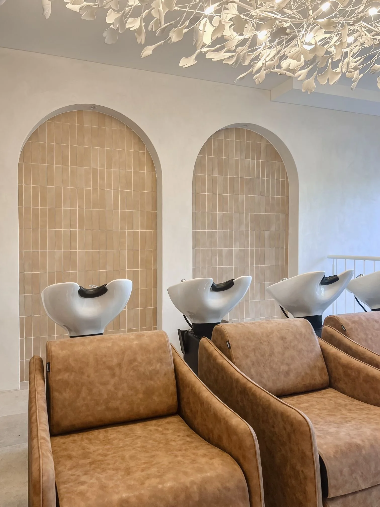 Salon chairs in a row facing hair washing stations with white sinks and beige chairs. A decorative white floral chandelier hangs above. The wall features beige tiled arches.
