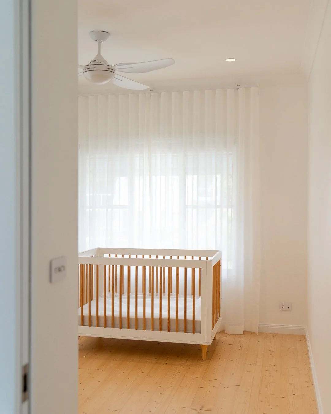 A minimalist nursery with a white crib, sheer white curtains, a ceiling fan, and hardwood floors.