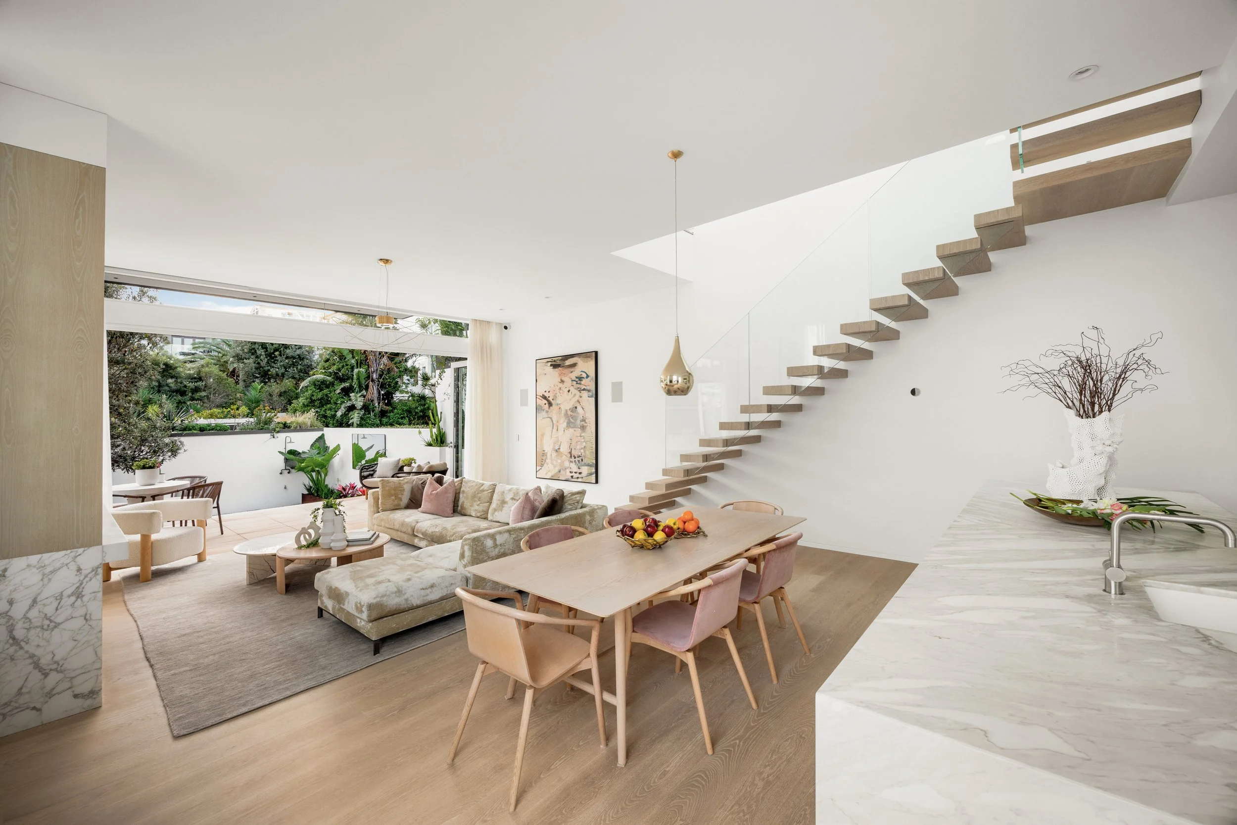 Modern open-plan living space with beige and white decor, a dining table with chairs, a sofa, and a view of an outdoor patio with greenery through large windows.