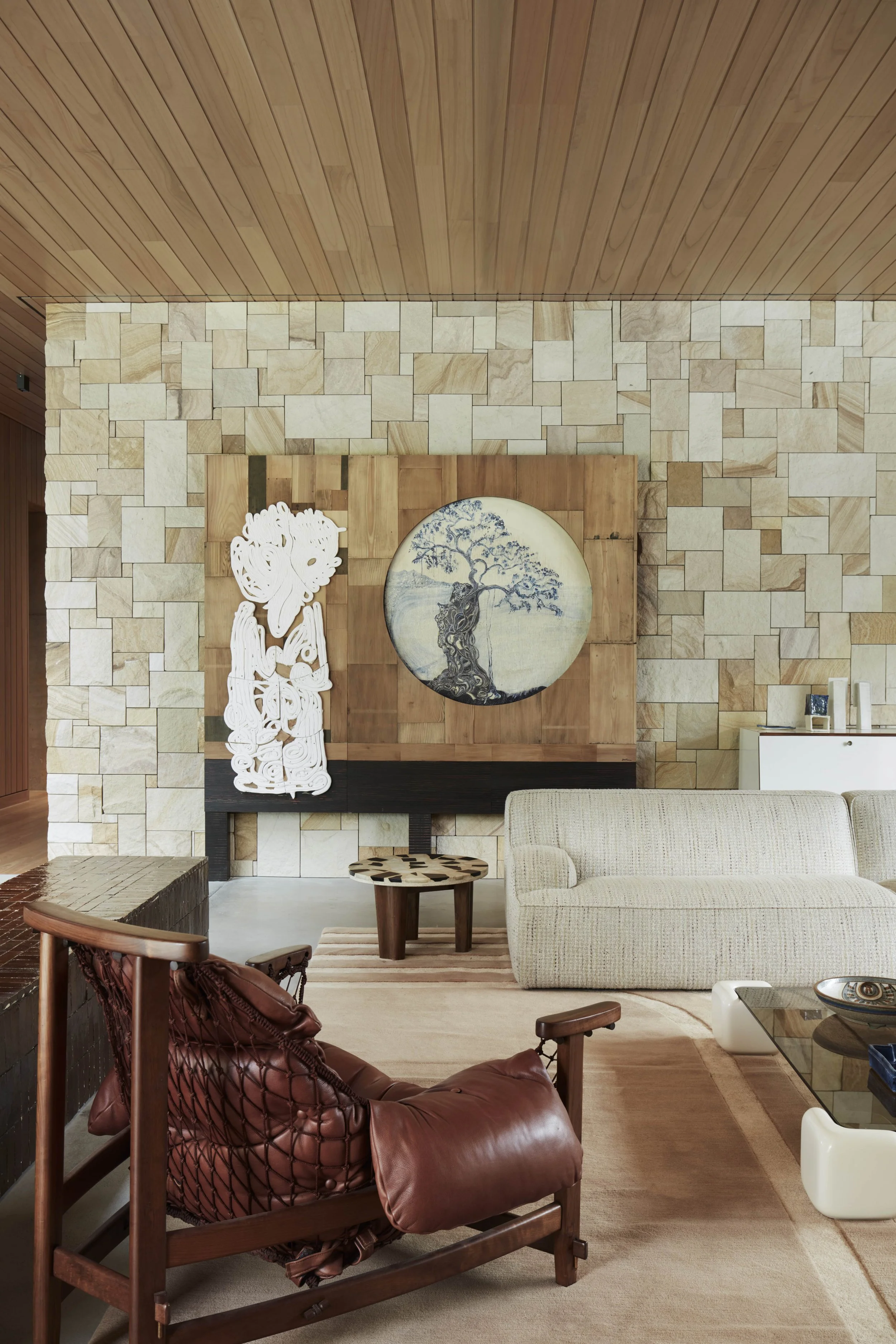 Living room with textured brick wall, wooden ceiling, artwork of a tree on a round canvas, and various furniture including a white sofa, a wooden chair, and a glass coffee table.