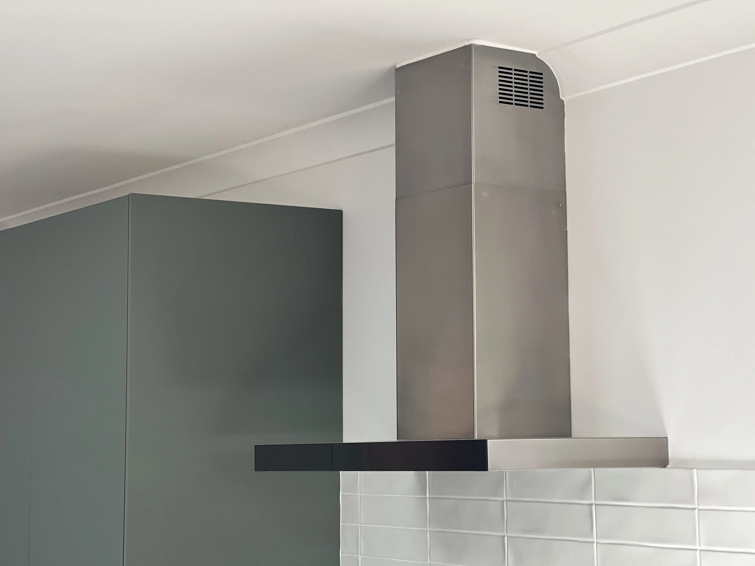 A bathroom corner with a gray wall and a section of white tiled wall below, a dark gray cabinet, and a stainless steel vent pipe extending from the ceiling.