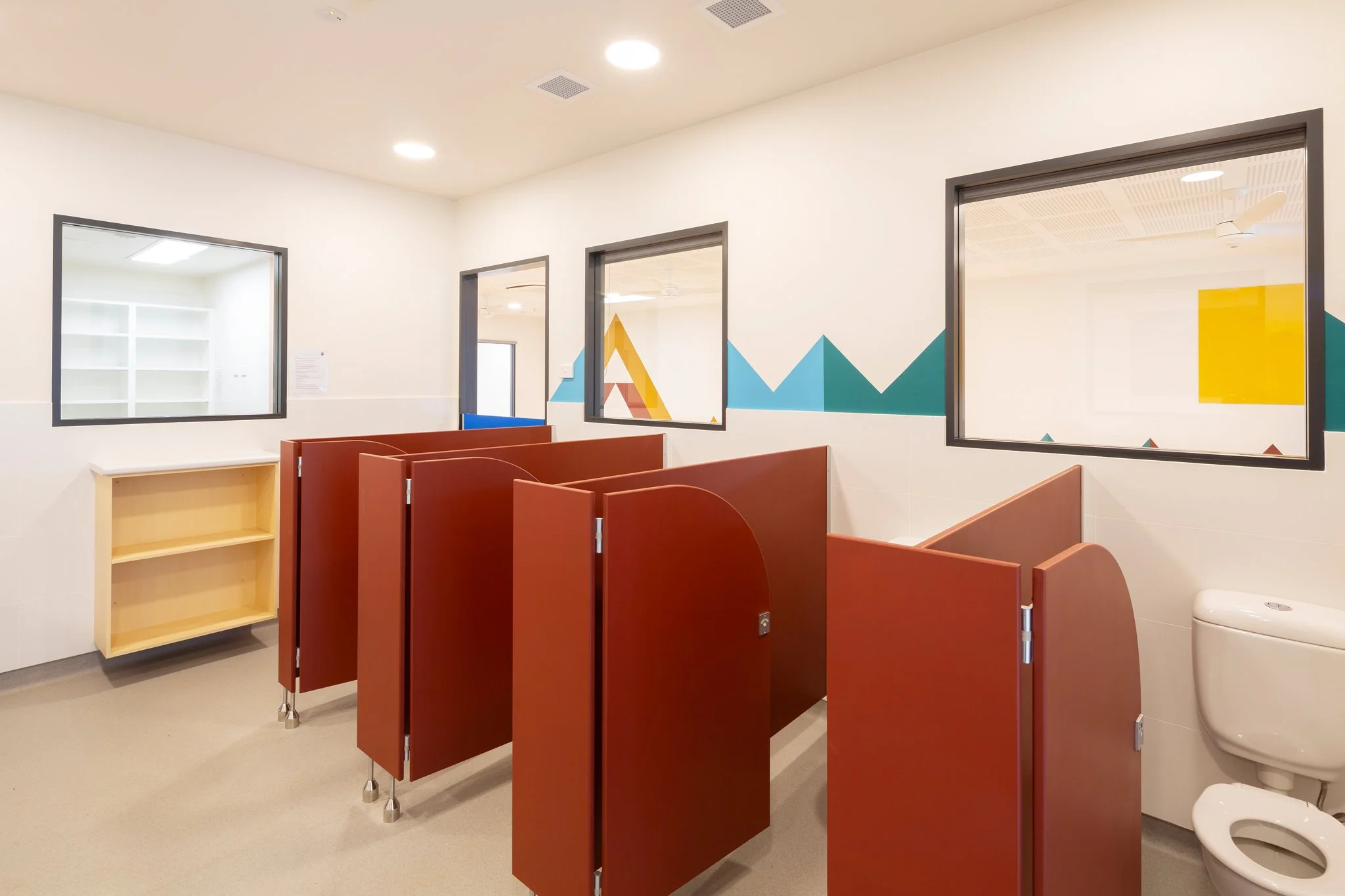 A restroom with multiple stalls, a window, and a colorful mountain-themed wall art. There is a wooden shelf and a toilet visible.