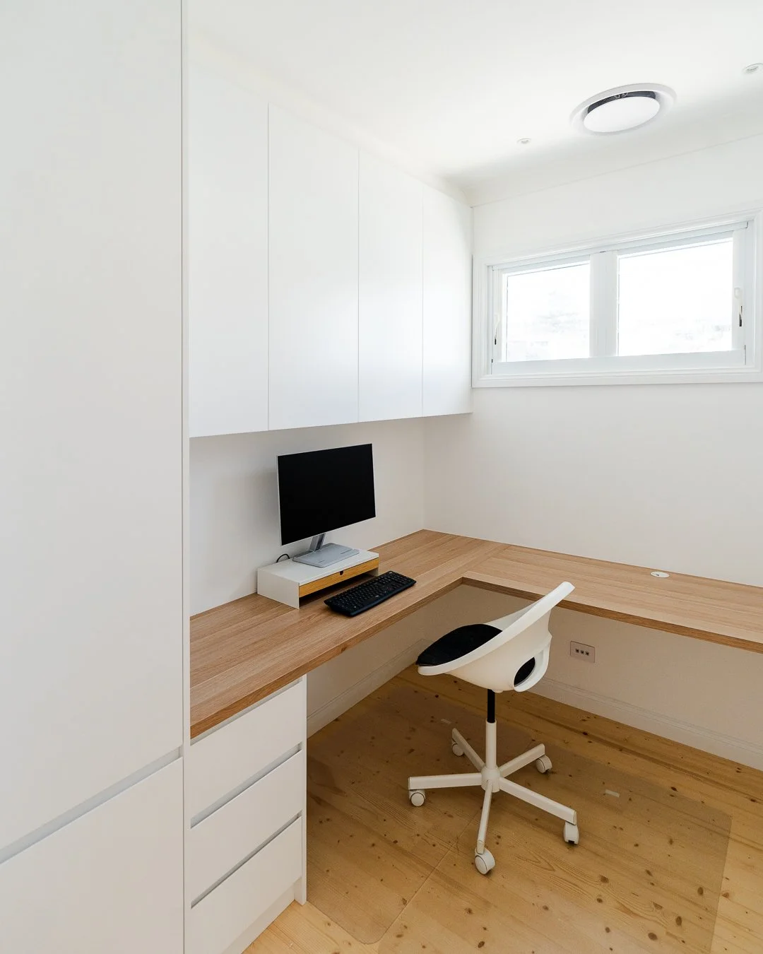 Minimalist home office with a wooden desk, a white swivel chair, a small desktop computer, and white cabinetry, illuminated by natural light from a window.
