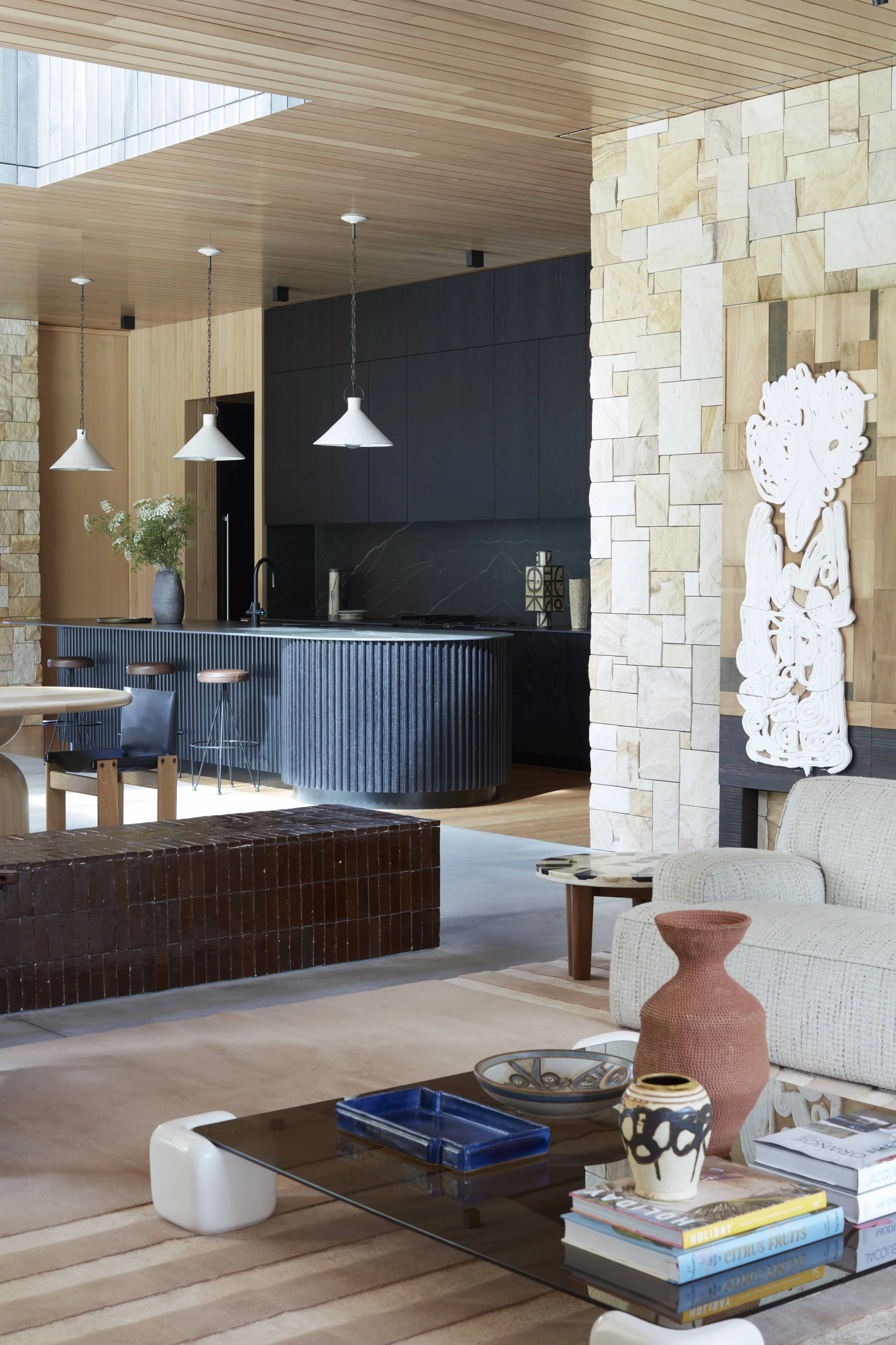 Interior of a modern living space featuring a kitchen with black cabinetry, a stone accent wall, and a wooden ceiling. There is a white sculpture on the wall, a beige sofa with a small round table, and a glass coffee table with decorative vases and b
