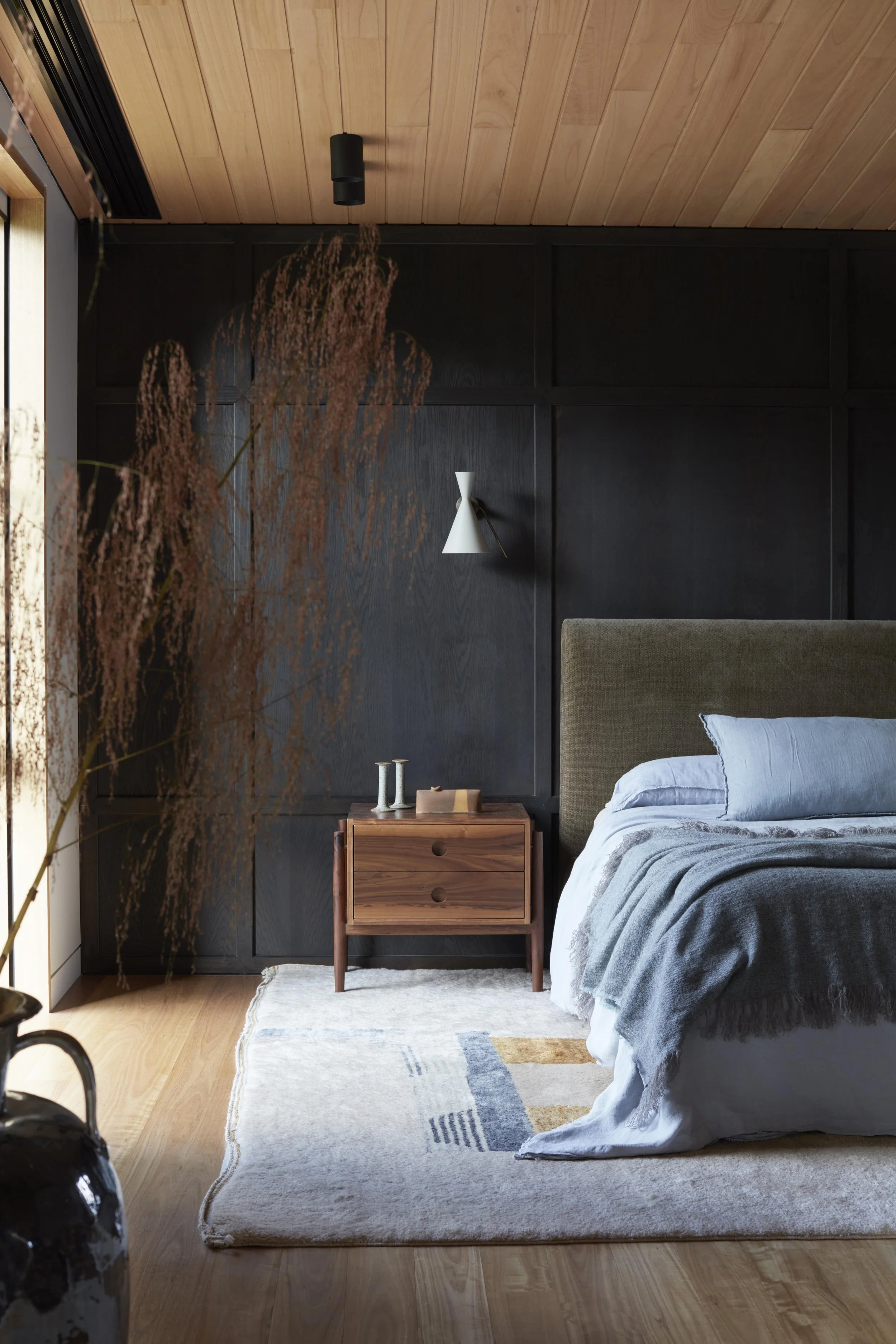 A bedroom with a gray bed and bedding, a wooden nightstand with decorative items, dark paneled wall with a white wall sconce, pale wood ceiling, and a large light-colored area rug with geometric patterns.
