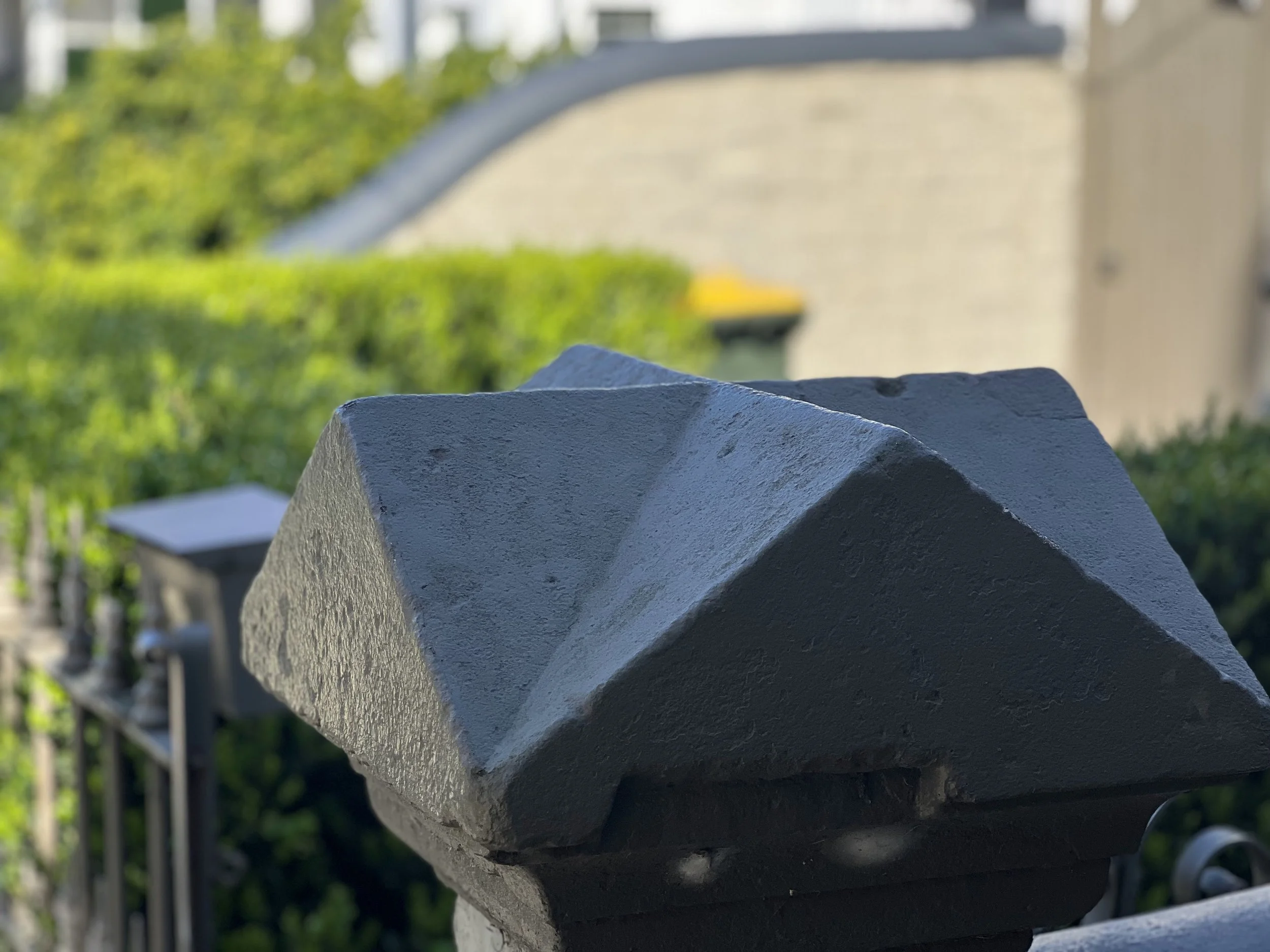 Close-up view of a row of gray painted metal or concrete beams or bars with a textured surface, with a blurred background of green foliage and a building.