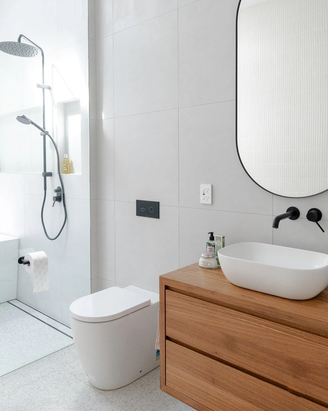Modern bathroom with walk-in shower, wall-mounted toilet, and wooden vanity with oval vessel sink and black fixtures.