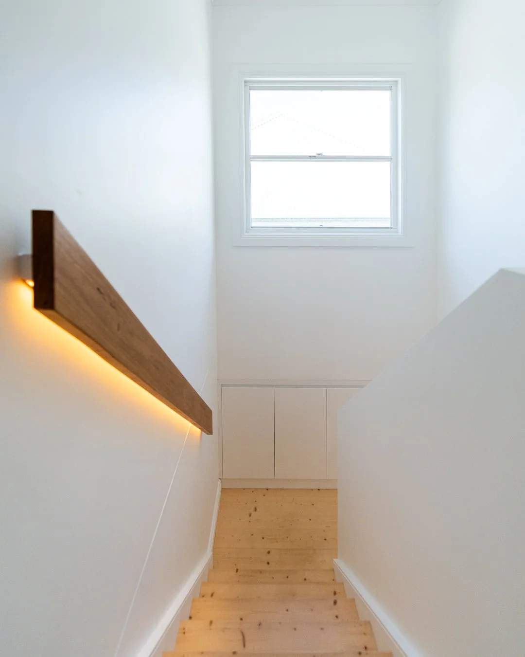 Top of a staircase with wooden steps, white walls, a window at the top, and a wooden handrail with LED lighting.