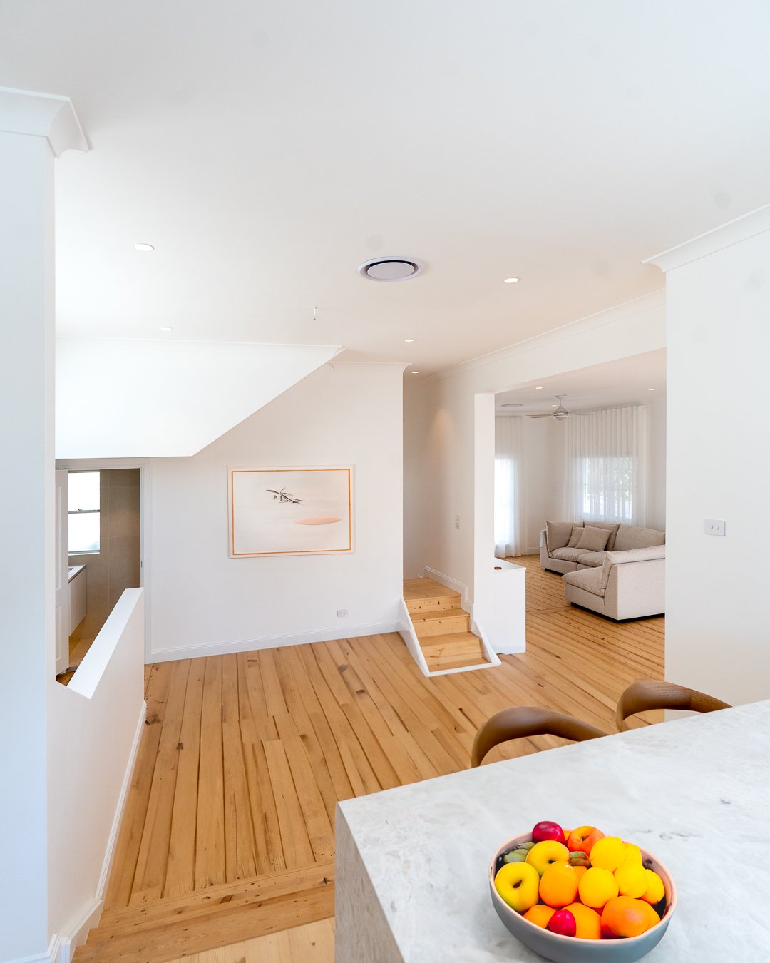 Bright, spacious living room with white walls, wooden floors, and natural light, featuring a sitting area with a beige couch and large windows with sheer curtains.