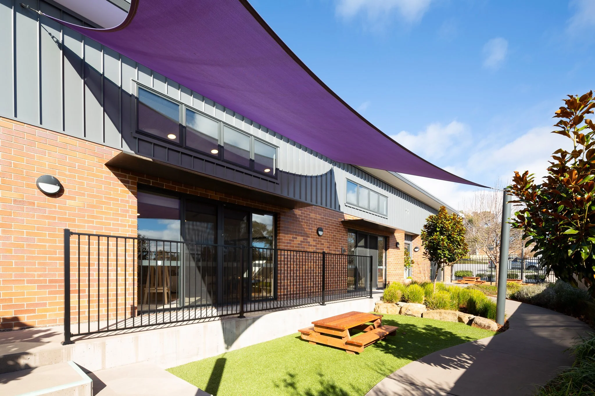 Exterior view of modern residential buildings with large windows, brick and metal siding, purple sunshade, fenced backyard with artificial grass, picnic table, and trees under a blue sky.