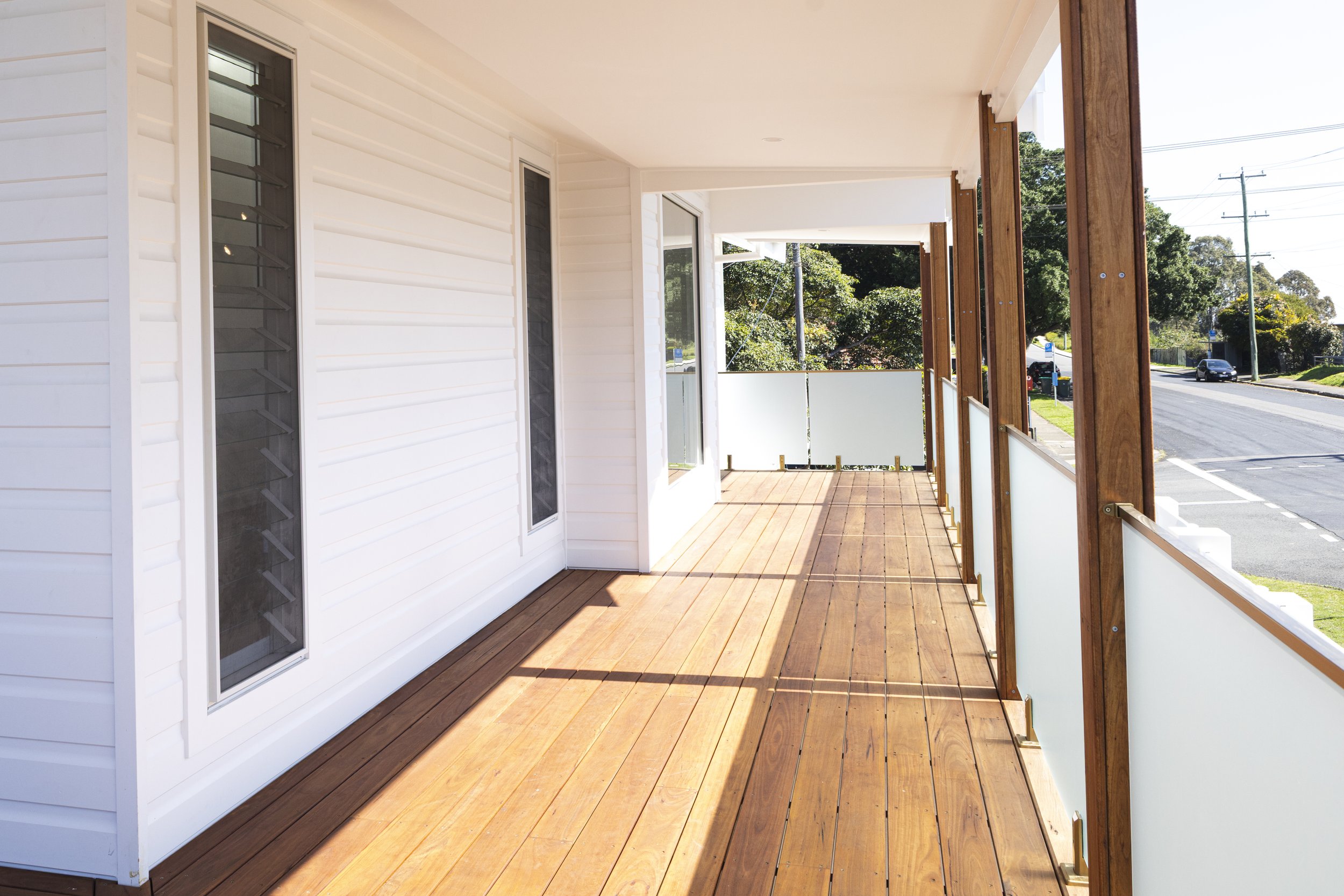 A view of a porch with wooden flooring, white siding exterior walls, and a railing with frosted glass panels. There are trees and a street visible in the background.