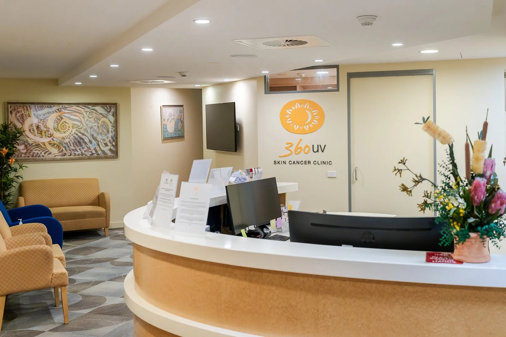 Reception area of a skin cancer clinic with a curved desk, computer monitors, informational pamphlets, and a floral arrangement. There are chairs and artwork on the walls, with the clinic's logo on the wall that says '360 UV Skin Cancer Clinic'.