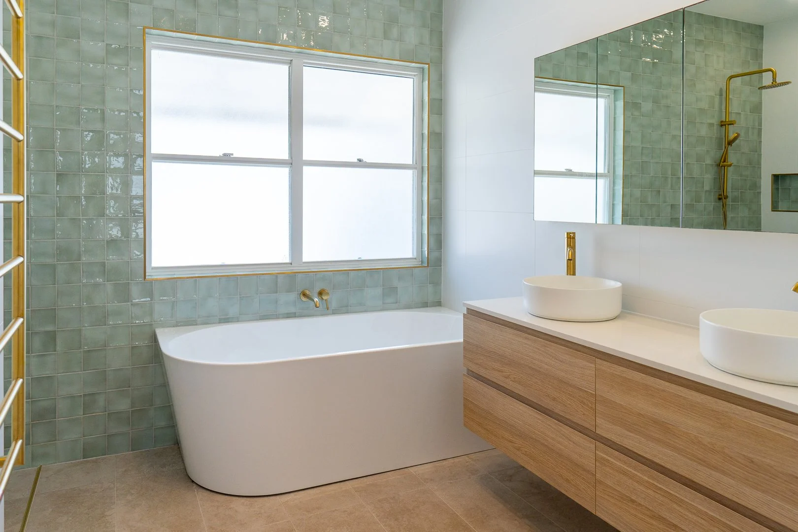 Modern bathroom with a white bathtub, green tiled walls, large window, double vessel sinks on a wooden vanity, gold fixtures, and a large mirror.