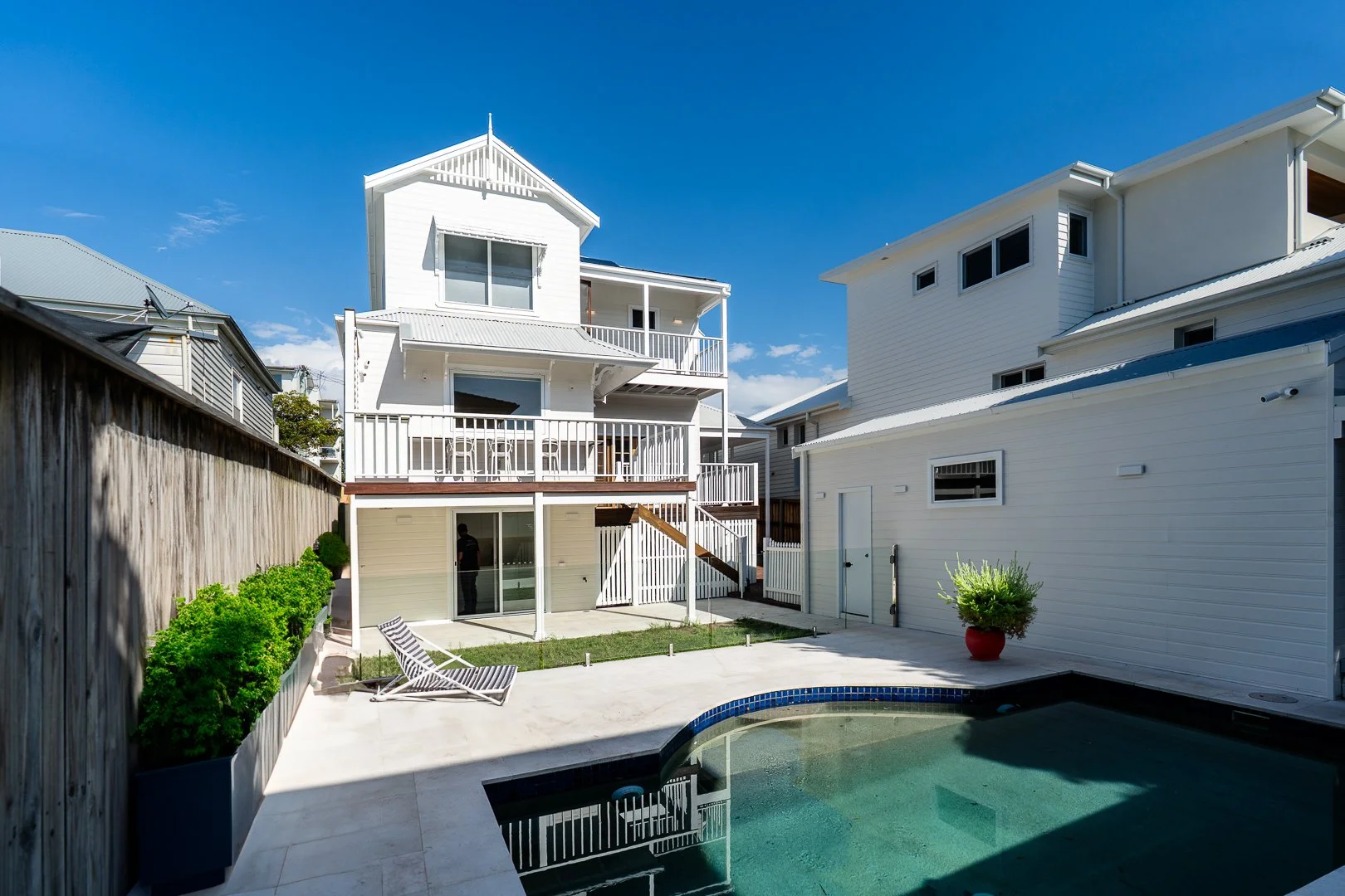 Backyard of a white multi-story house with balconies, a swimming pool, lounge chair, and potted plant under a blue sky.