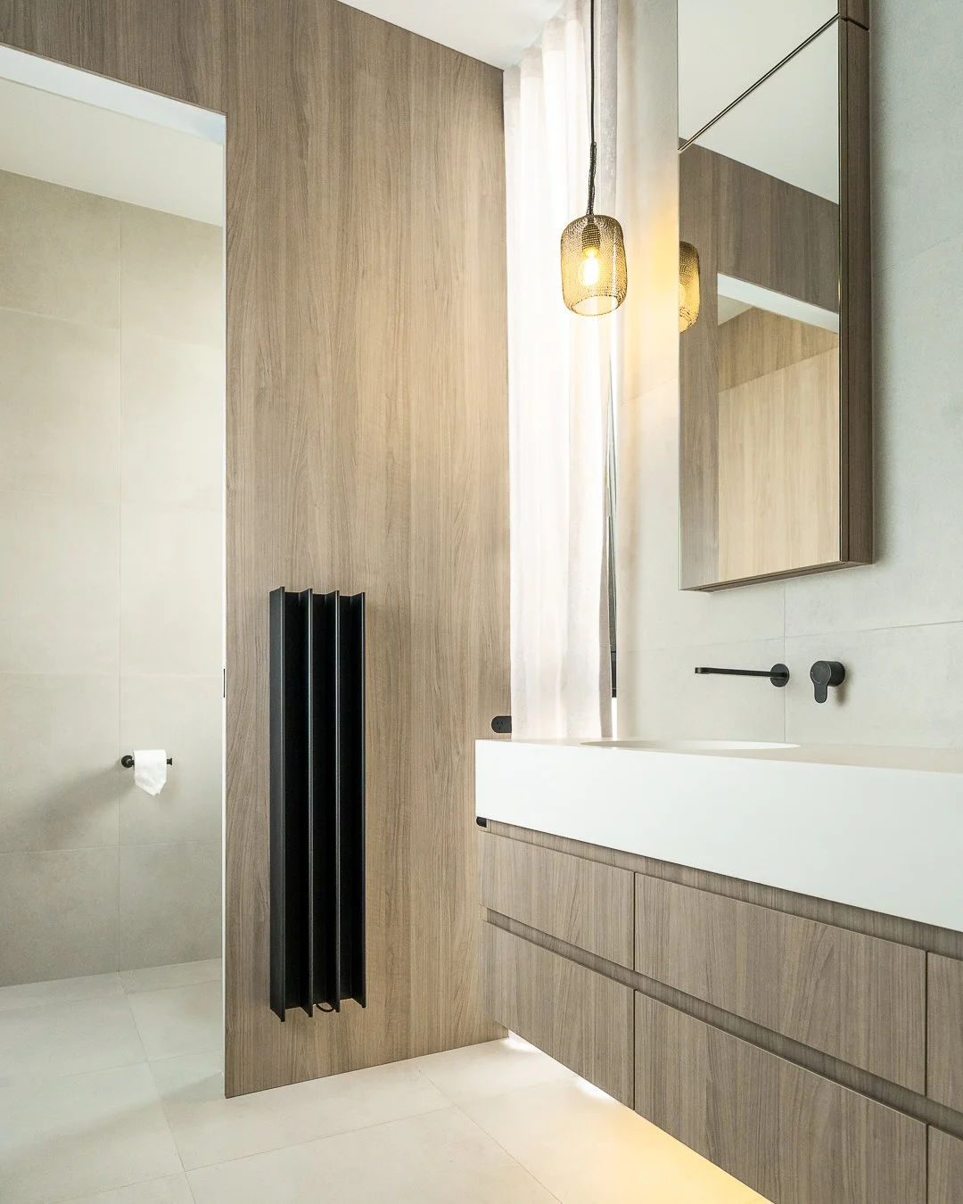 Modern bathroom with wooden cabinetry, a large mirror, black fixtures, black radiator, and a hanging light fixture.