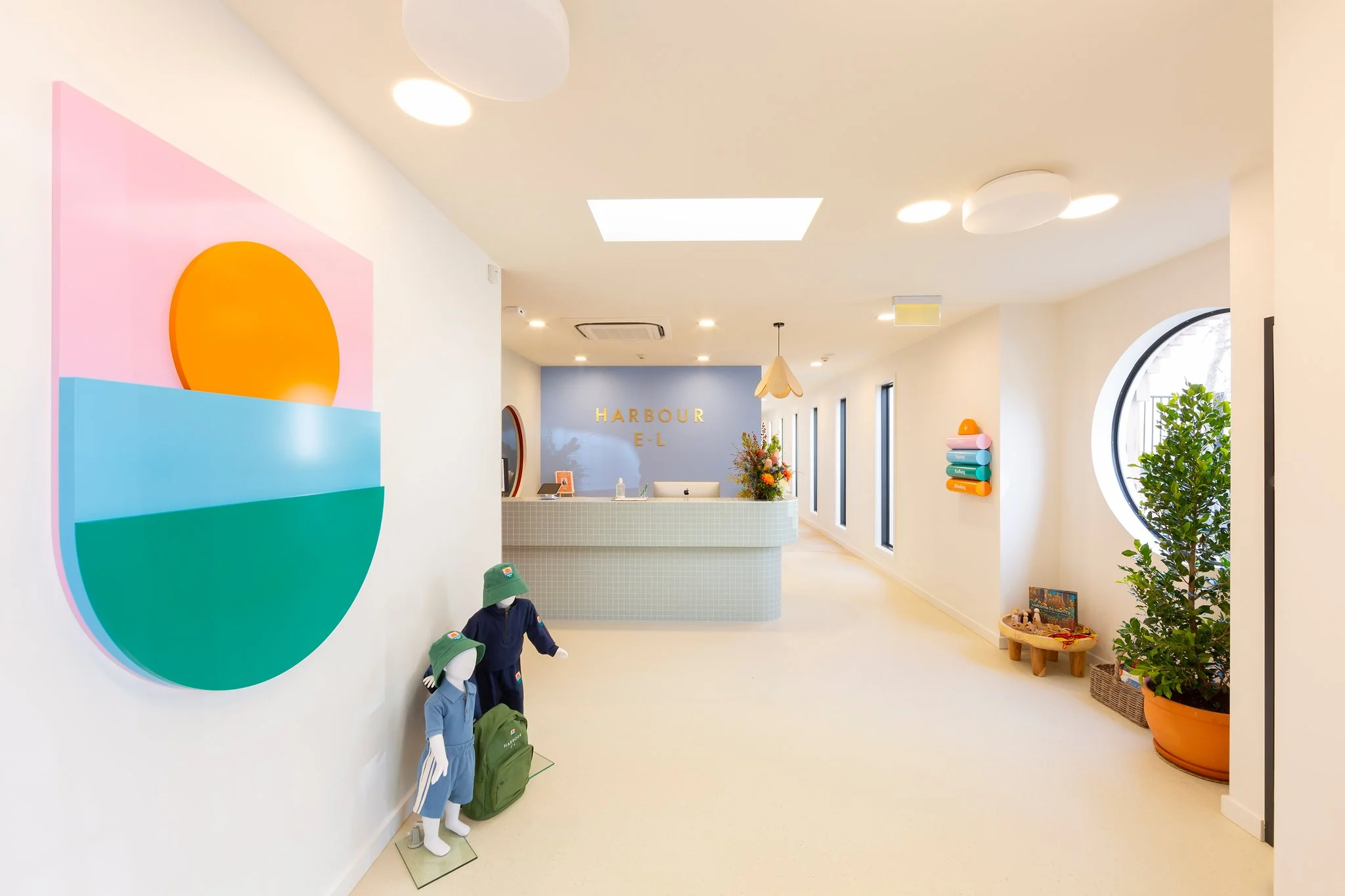 The image shows the reception area of a modern, brightly lit establishment with colorful decor. On the left wall, there is a large, abstract artwork featuring semi-circular shapes in pink, orange, blue, and green. In front of the wall, three mannequi