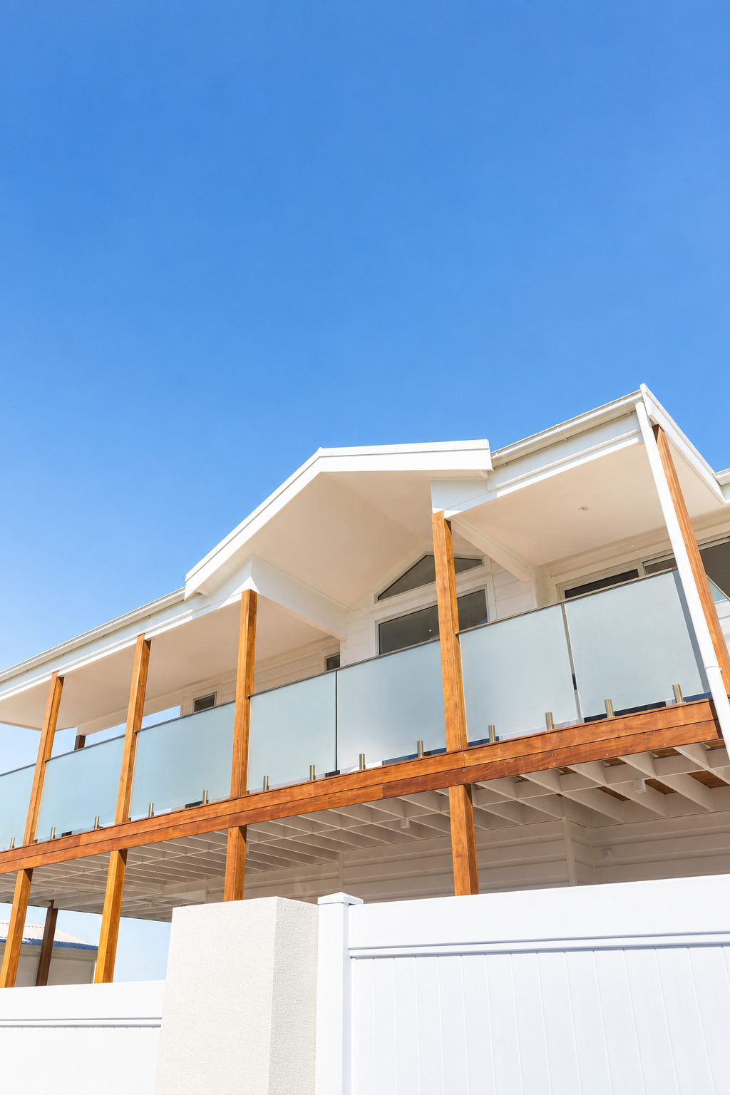 A modern house with a white facade and wooden accents, featuring a balcony with glass railings, under a clear blue sky.