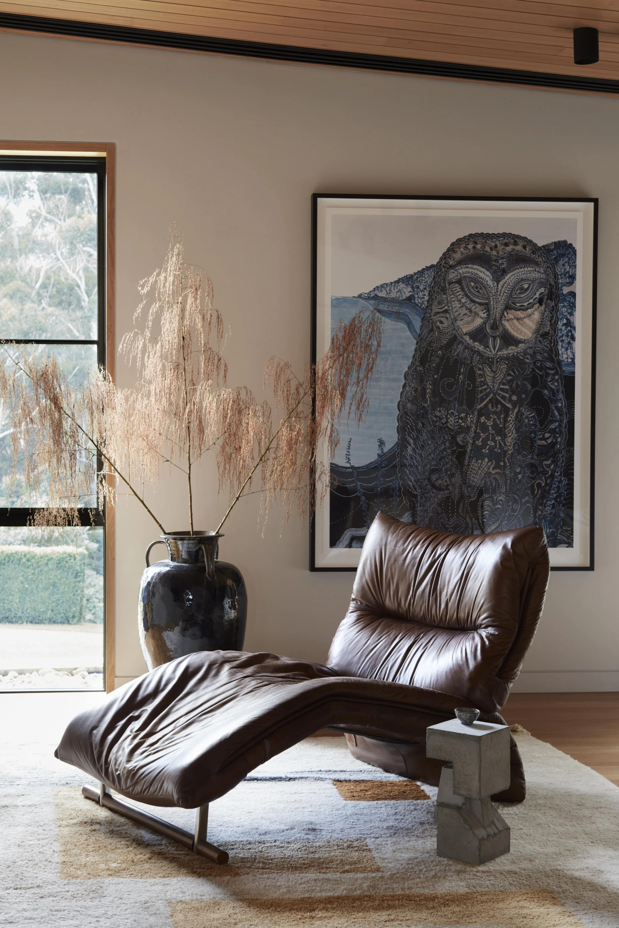 Modern living room with a brown leather lounge chair, a large decorative vase with dried branches, a picture of an owl on the wall, a window revealing a garden, and a beige rug with geometric pattern.