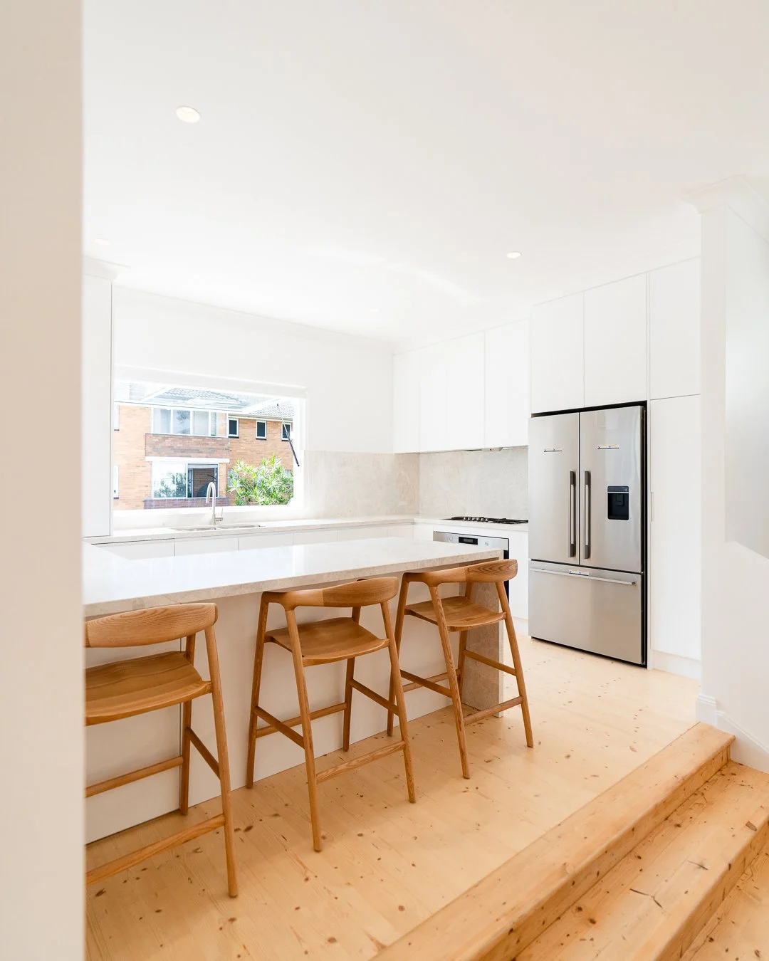 Modern kitchen with white cabinets, a marble countertop, a stainless steel refrigerator, and three wooden bar stools.