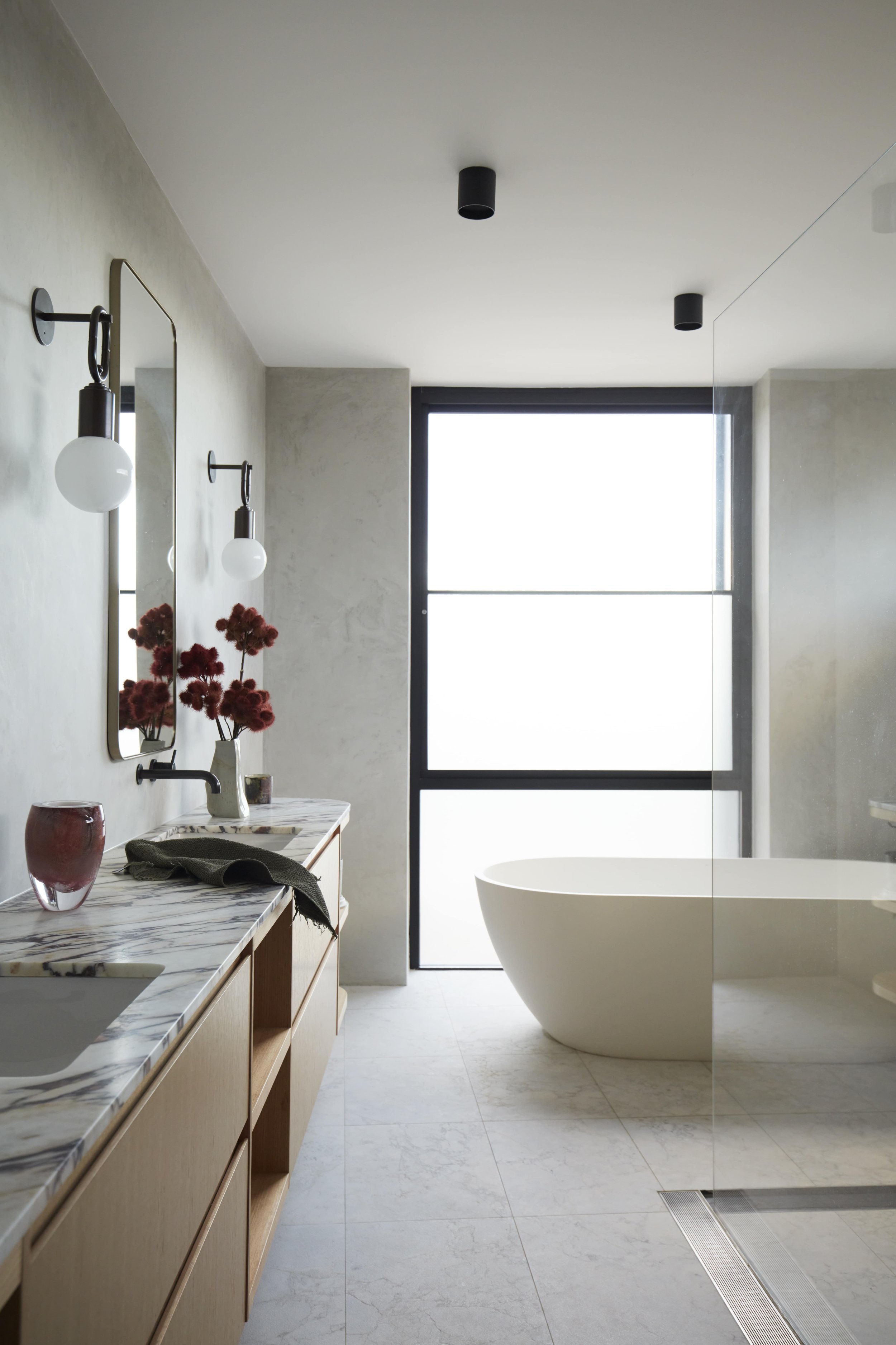 A modern bathroom with a freestanding bathtub, a large window, a marble countertop with a sink, two wall-mounted light fixtures, a tall mirror, and a vase with red flowers.
