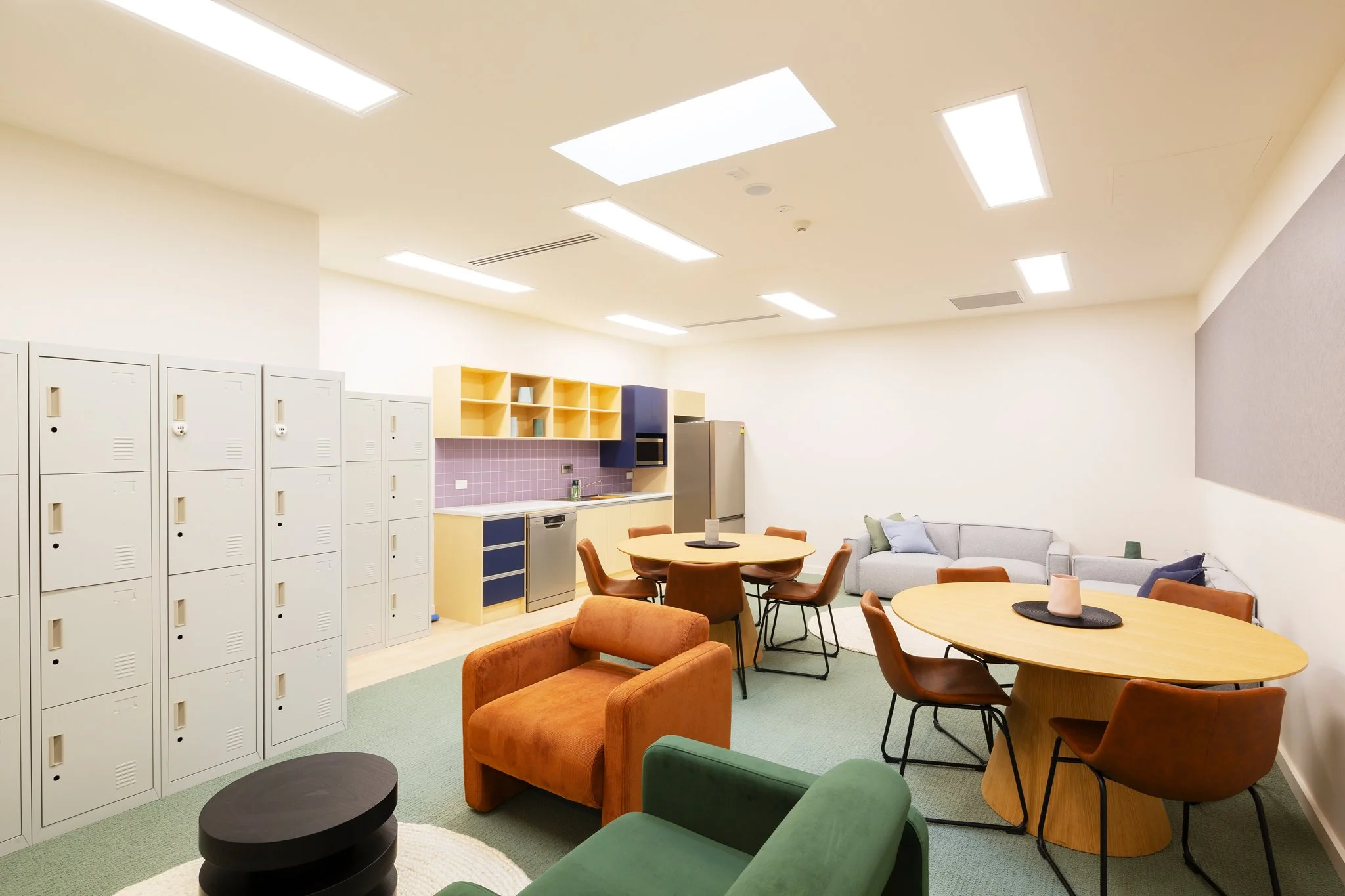 A modern break room with lockers, tables, colorful chairs, a small kitchen area, and a sitting area with a gray sofa and pillows.