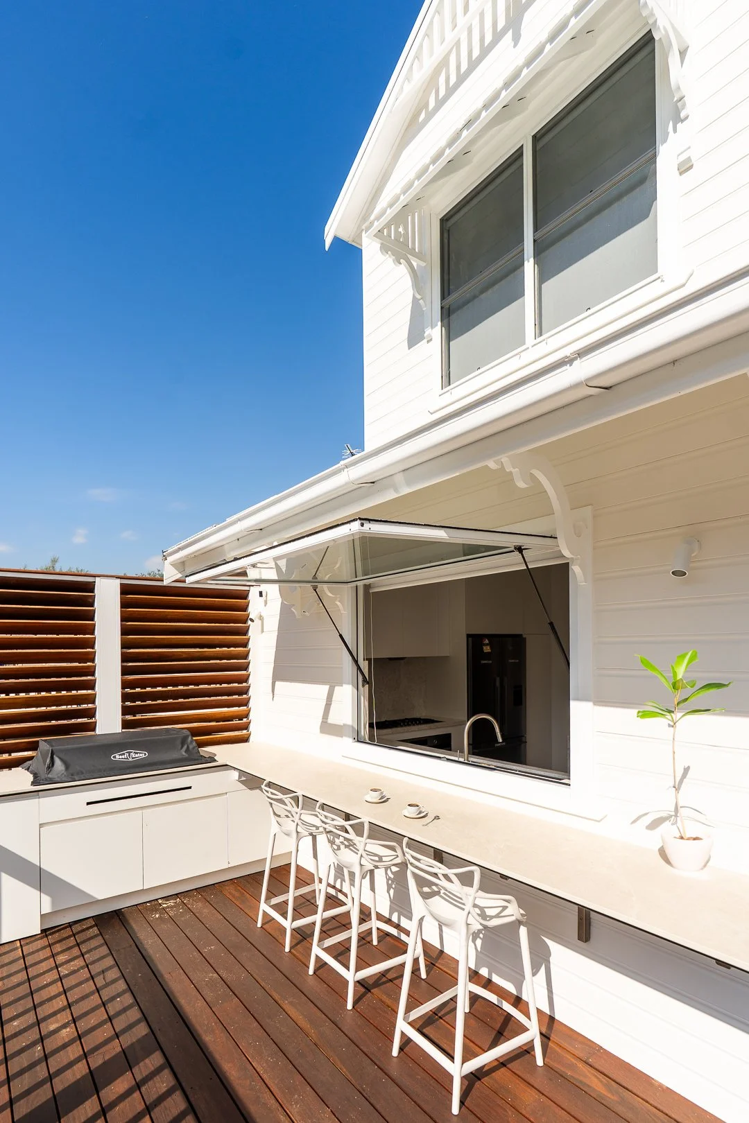 Outdoor patio area with white modern cabinetry, a built-in barbecue grill covered with a black cover, a long white counter with four white chairs, a potted plant, and a large open window with a glass cover. The house has white siding and a wooden pri