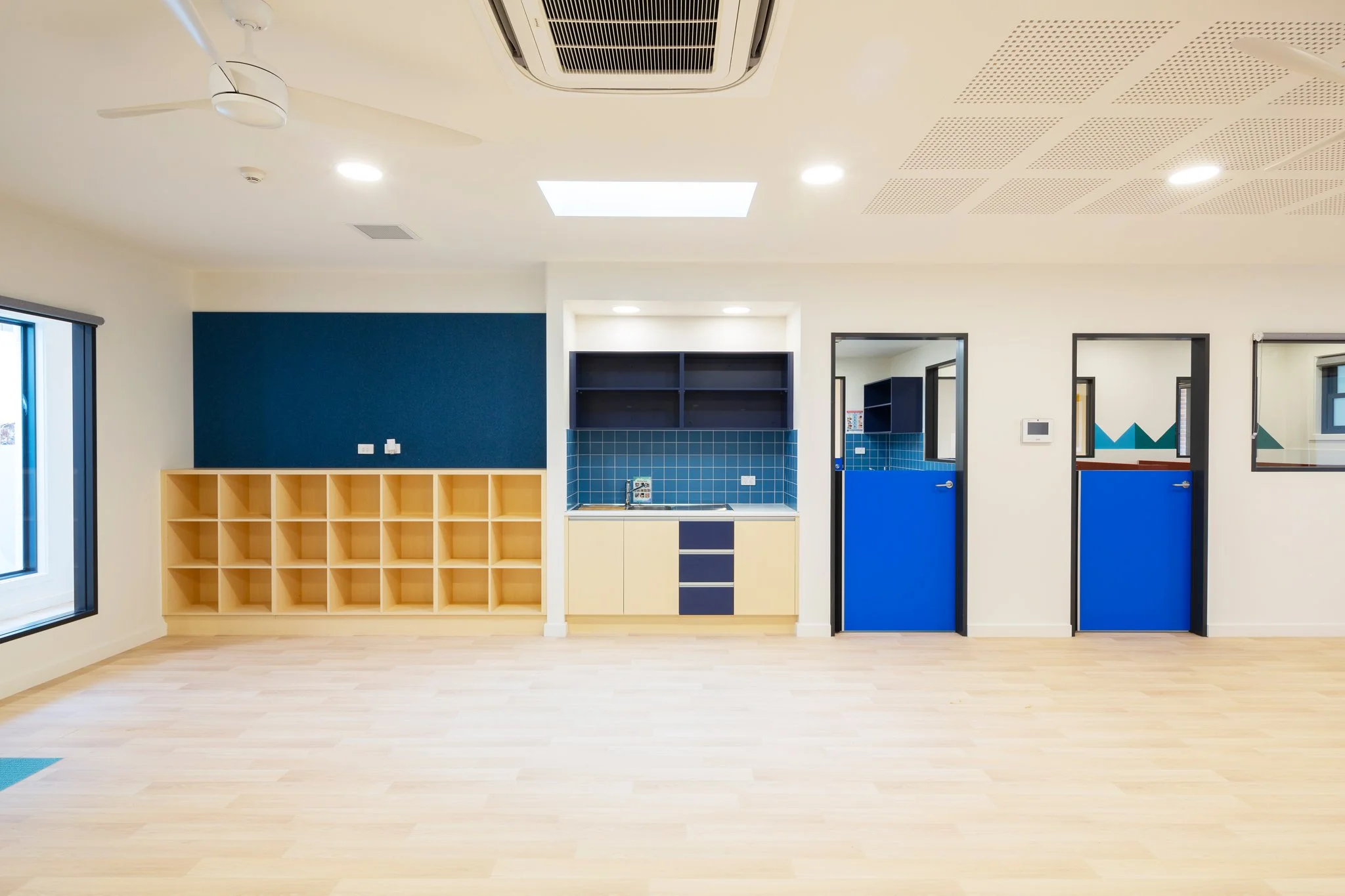 Empty classroom or daycare room with a blue and yellow storage unit, a kitchenette area with blue tiles, and blue doors. The ceiling has a white fan, air conditioning unit, and recessed lighting. The room has light-colored wooden floors and white wal