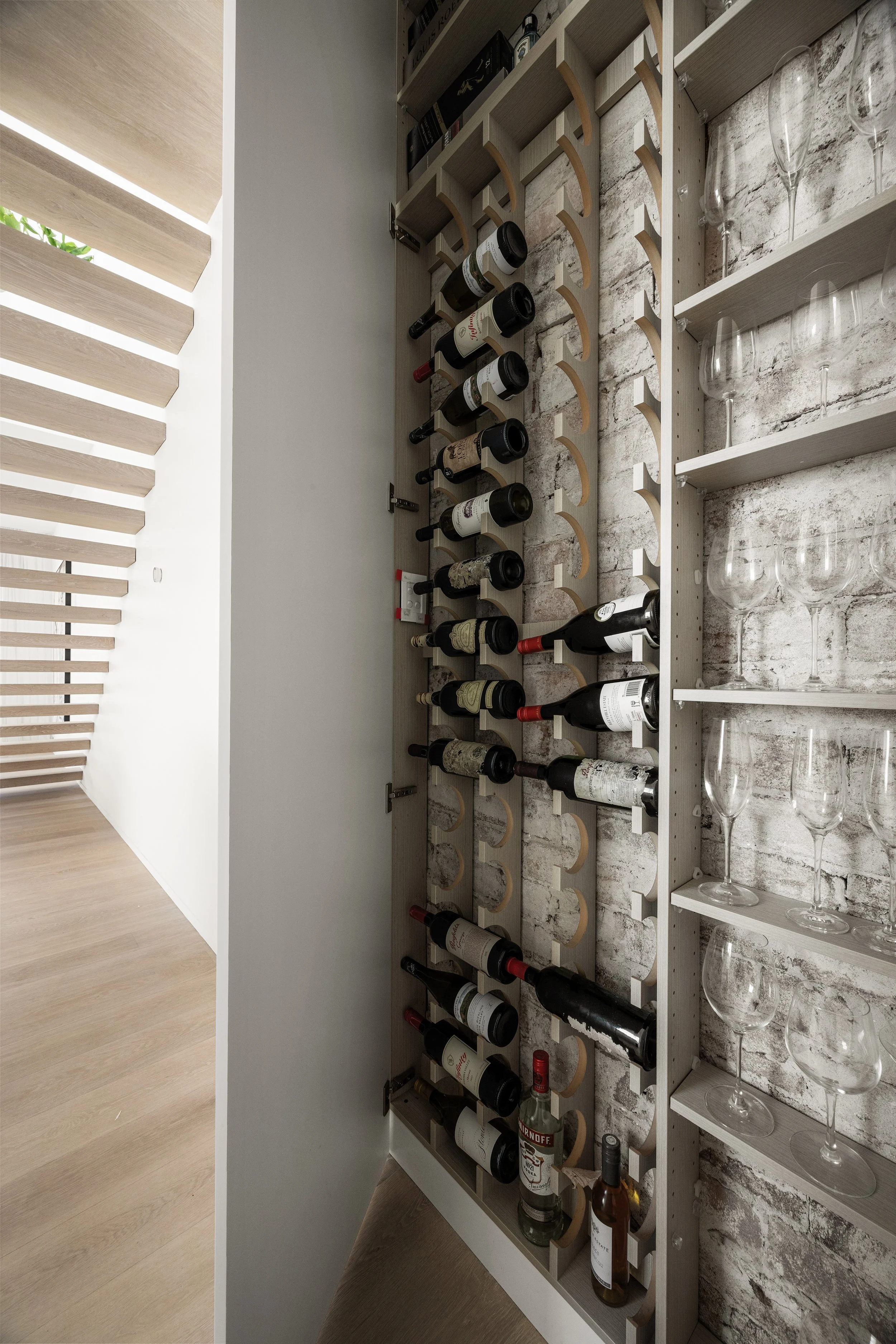 A wine storage shelf with bottles of wine, glasses, and a bottle of Smirnoff vodka, positioned against a white brick wall under a modern light-colored wooden ceiling with horizontal slats.