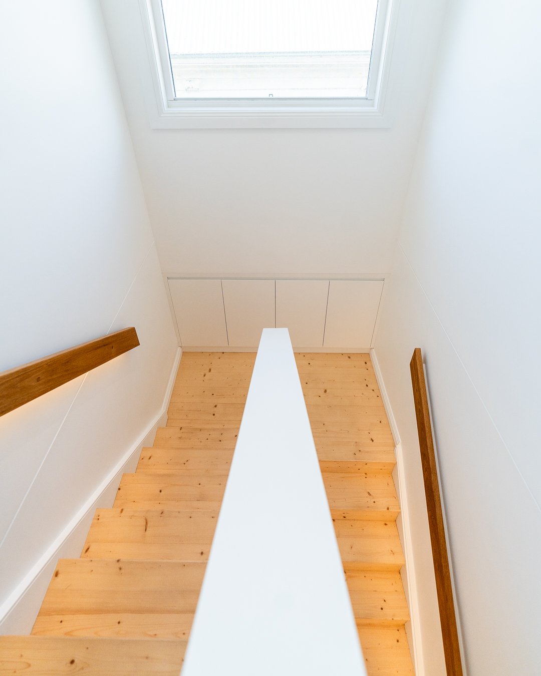 View looking down a staircase with wooden steps, white walls, a window at the top, and wooden handrails on both sides.