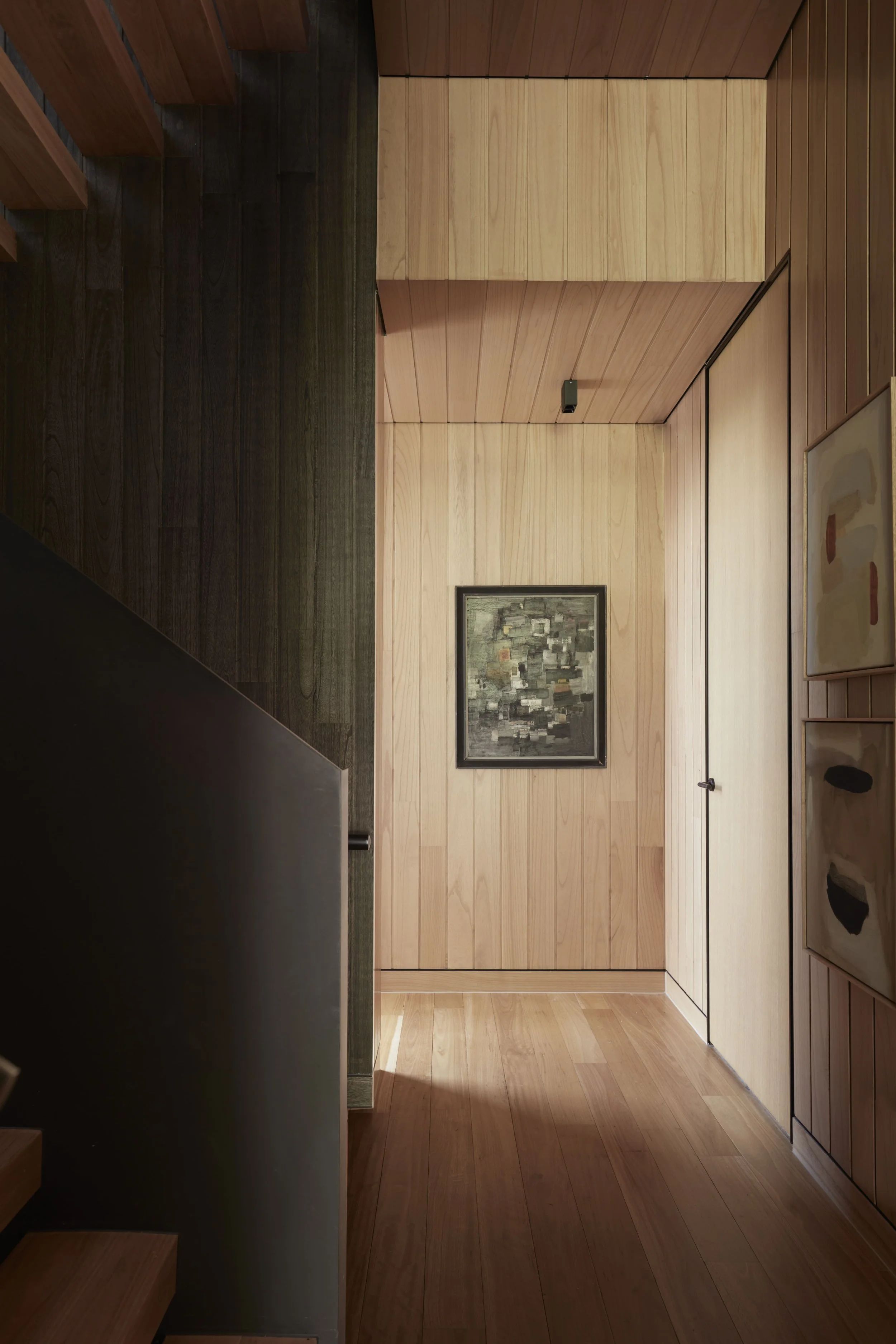 A hallway with wooden paneling on the walls and ceiling, featuring a framed abstract painting, a black speaker, and a black door handle.