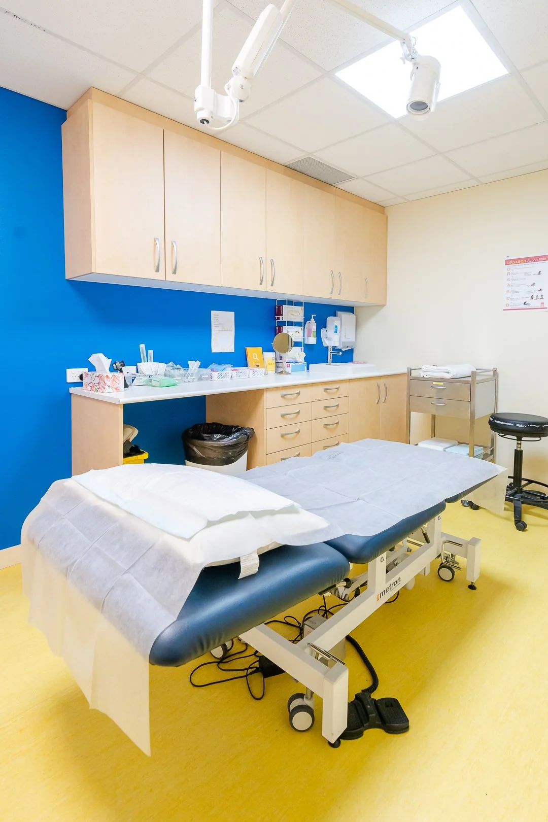 Empty medical examination room with an examination table covered by a paper sheet, blue and cream walls, overhead medical lights, and medical supplies on the counter.
