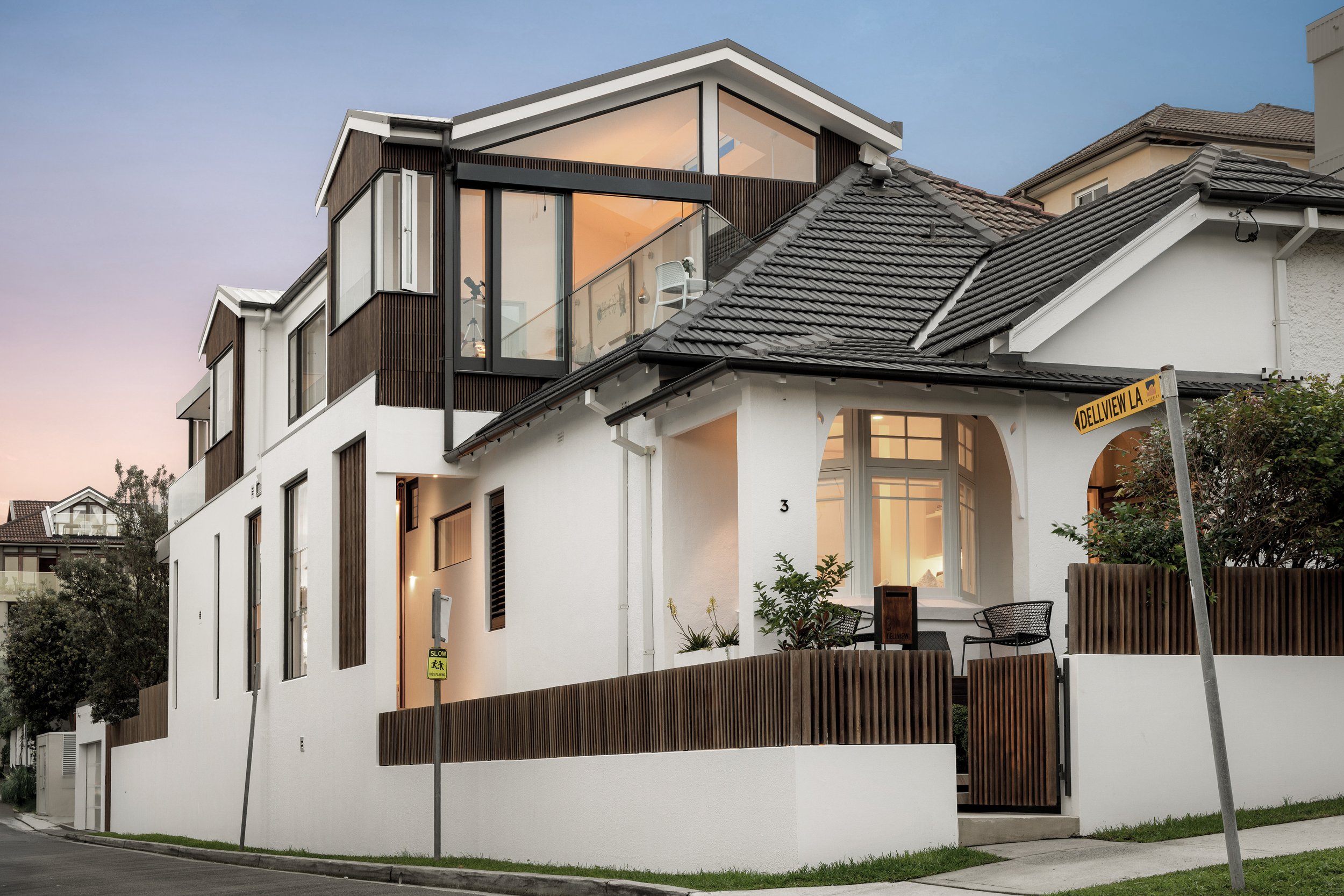 Modern multi-story house with white exterior walls, large glass windows, and a mix of flat and sloped roof sections. The house features wooden accents and is situated at the corner of a street with a fence and some greenery.